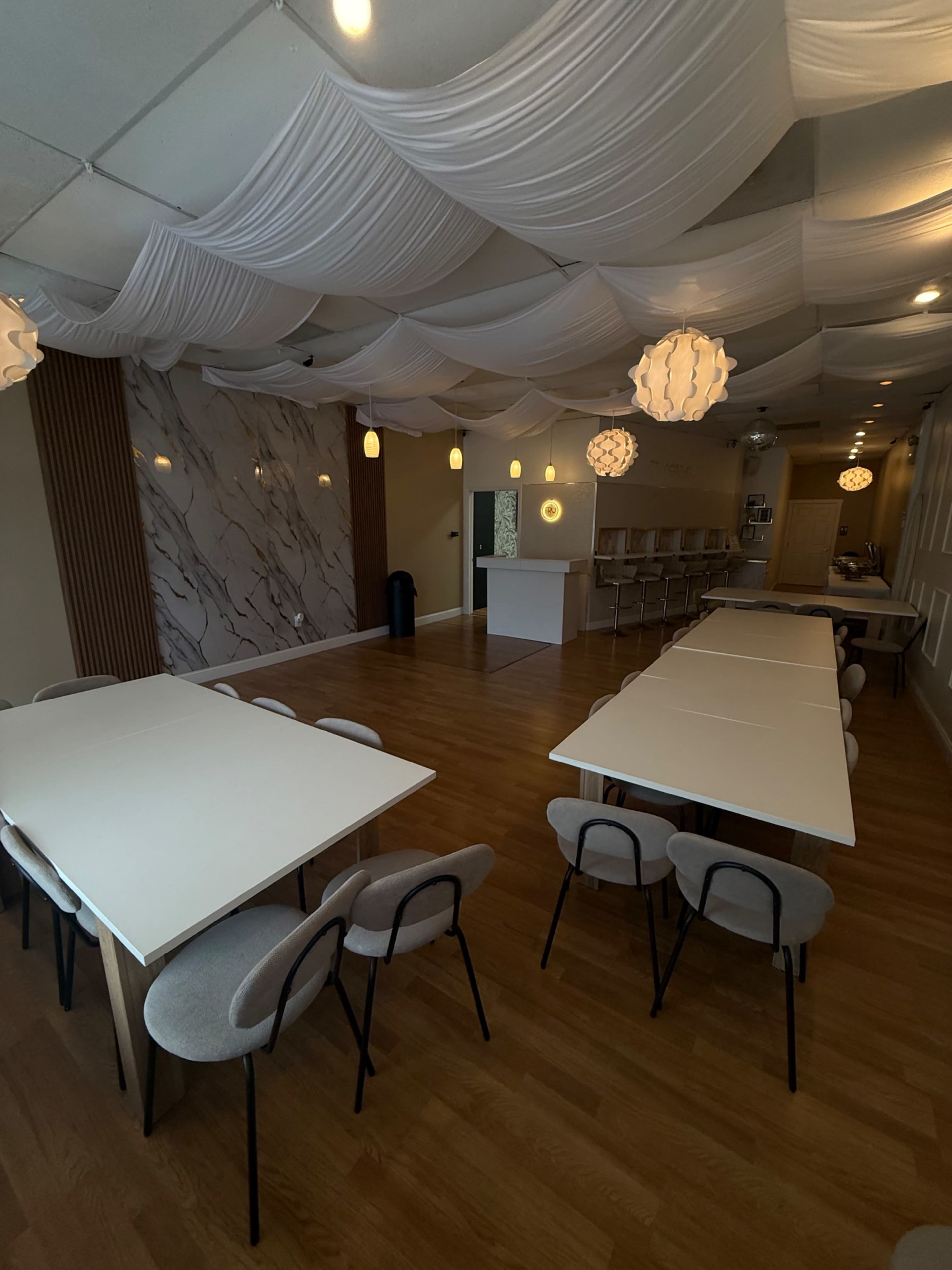 The image shows a spacious indoor venue with several tables arranged in a U-shape, draped fabric on the ceiling, and pendant lights hanging above.