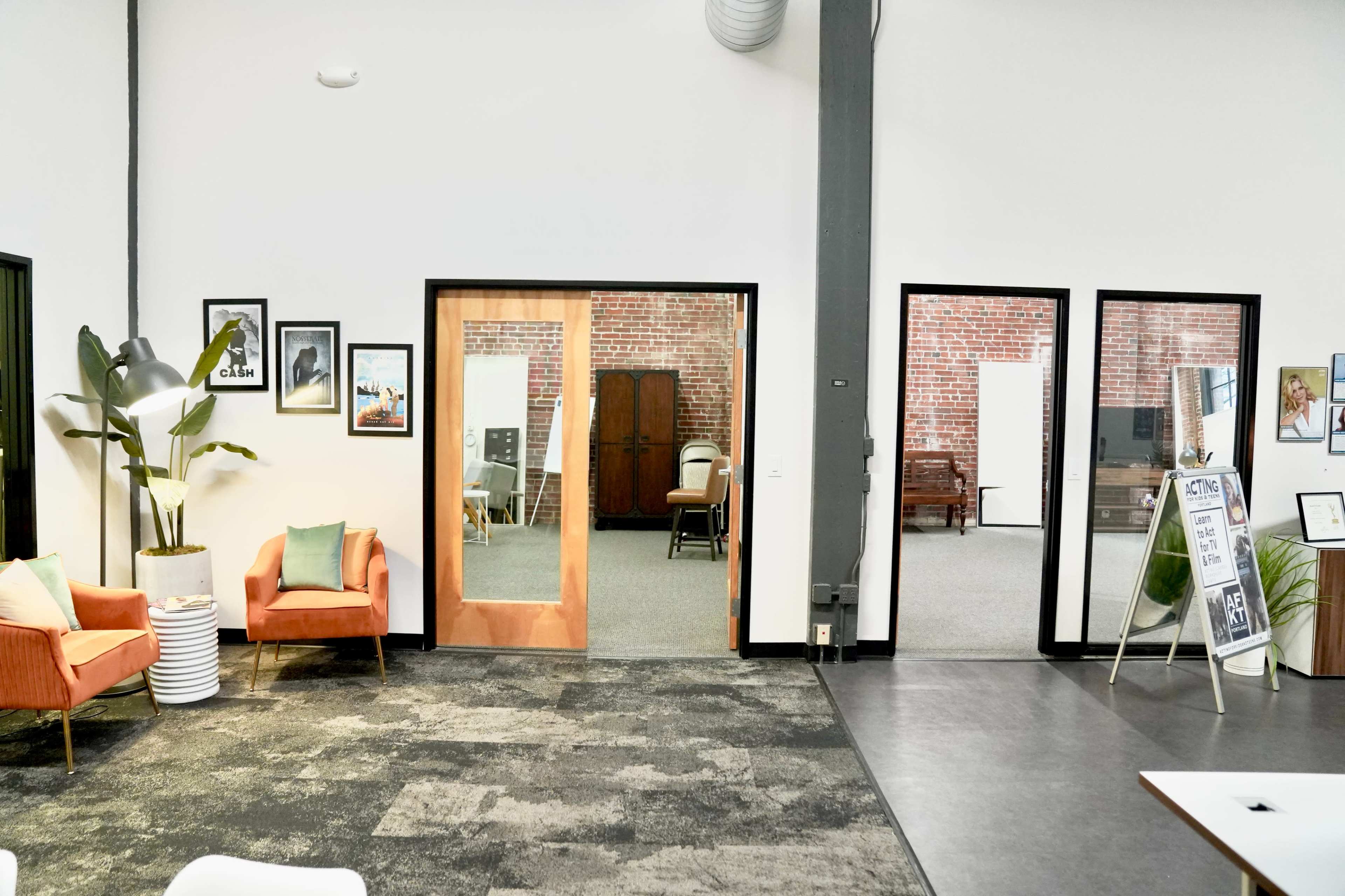 The image shows a modern office layout featuring a lounge area with orange chairs, two glass doors leading to another room, and a visible framed artwork on the walls.