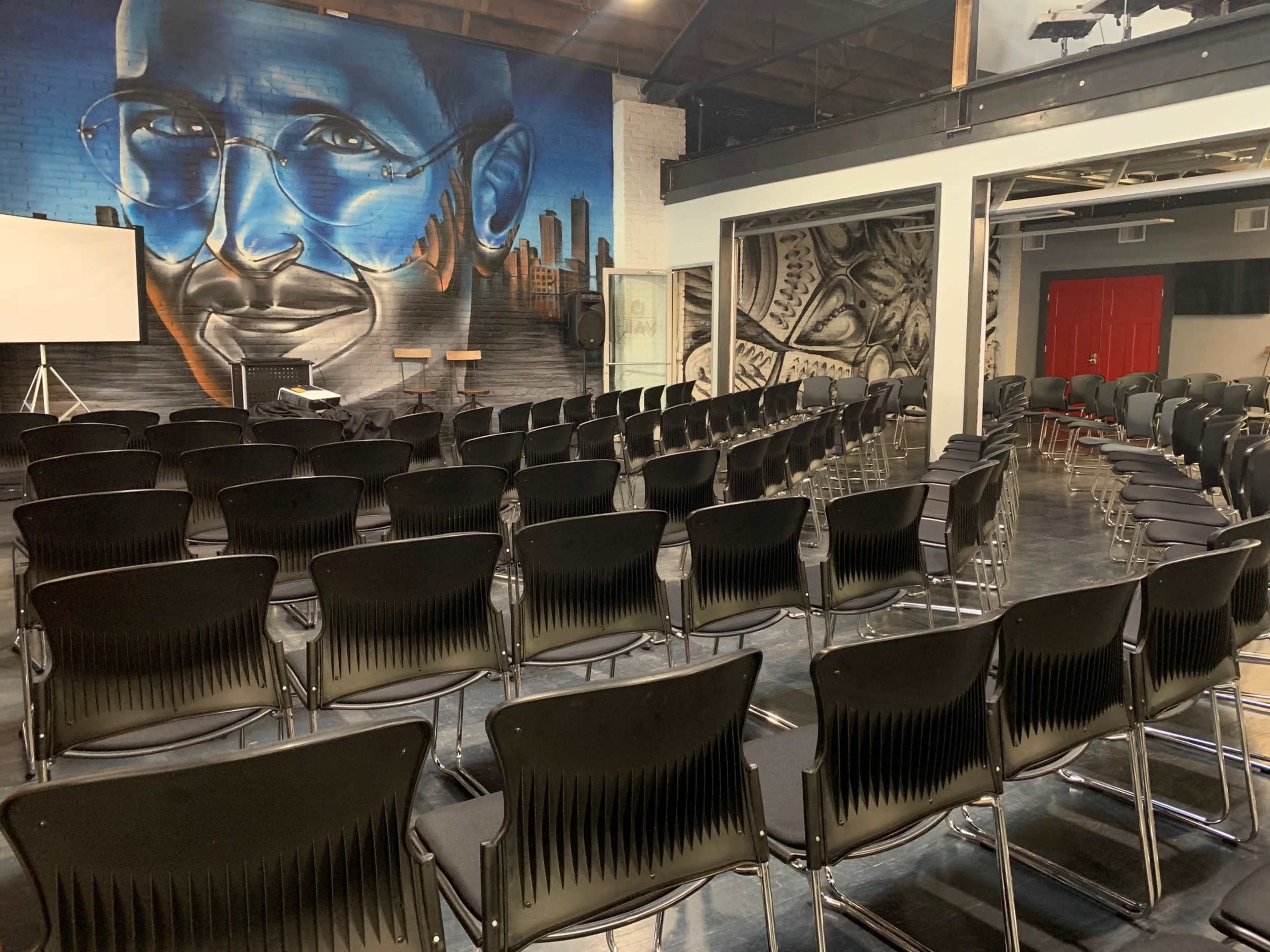 The image shows a spacious room set up with rows of black chairs facing a large mural of a man wearing glasses, with a projector screen visible at the front.