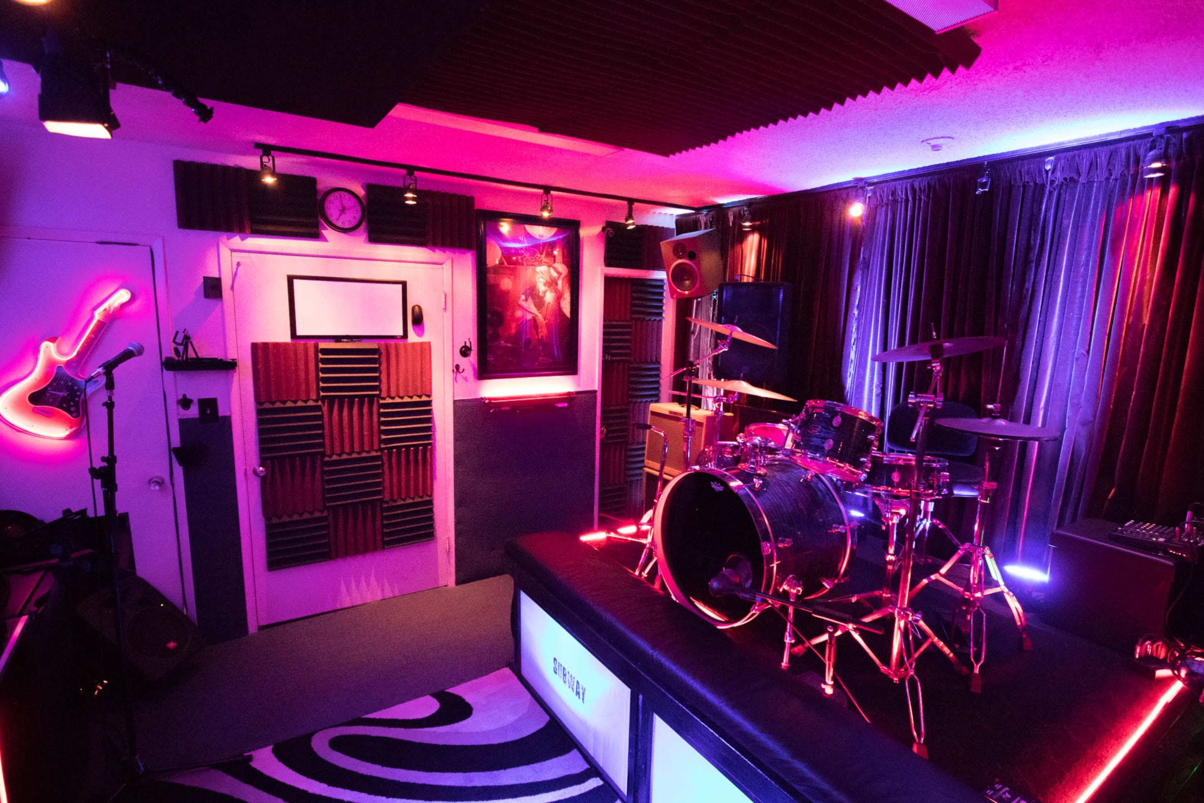The image shows a music rehearsal studio with a drum set, colorful lighting, soundproofing panels, and musical equipment arranged in a compact space.