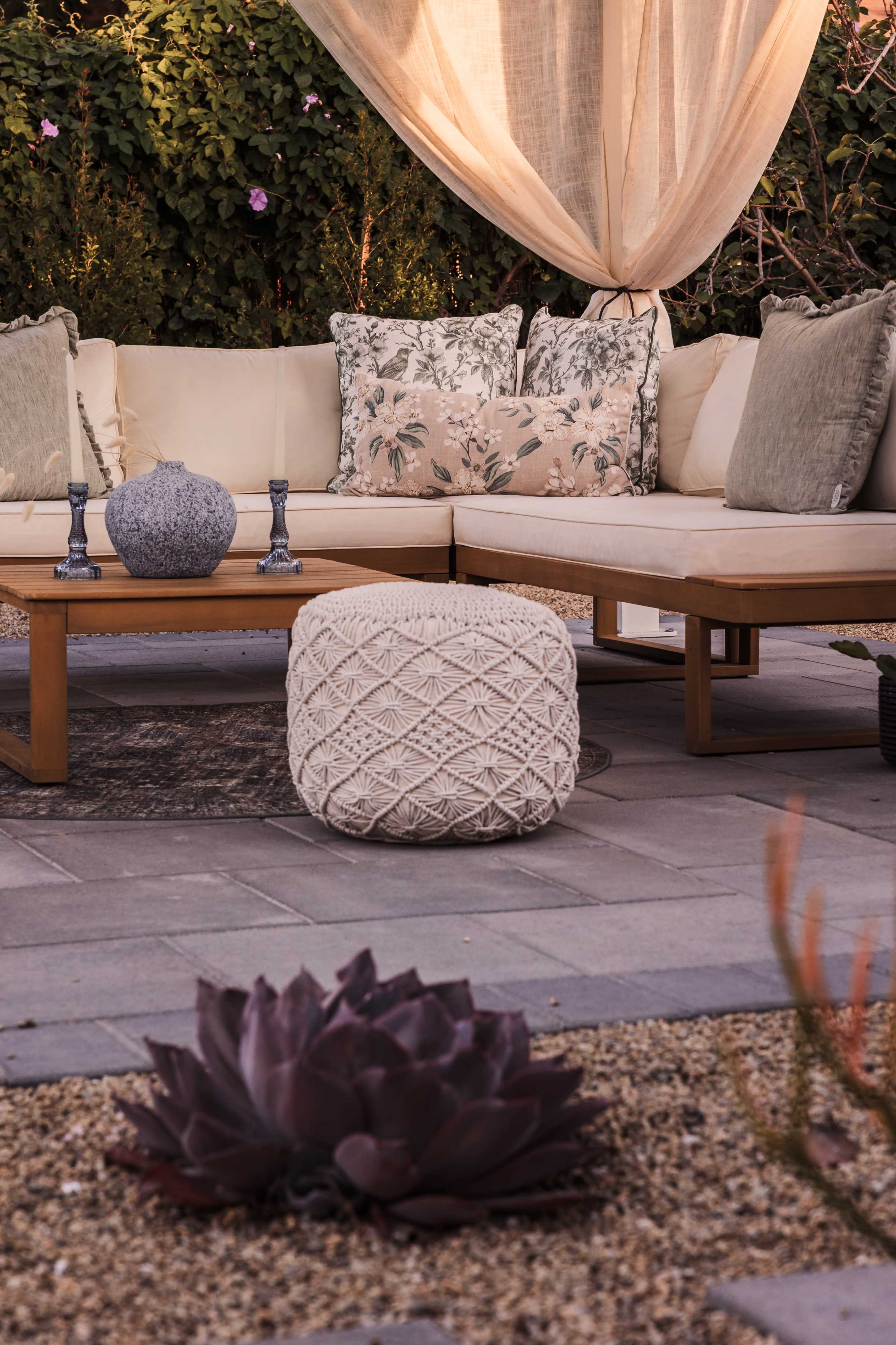 The scene features a cozy outdoor seating area with a large sectional sofa adorned with patterned cushions, a decorative pouf, and a succulent plant in a landscaped gravel setting.