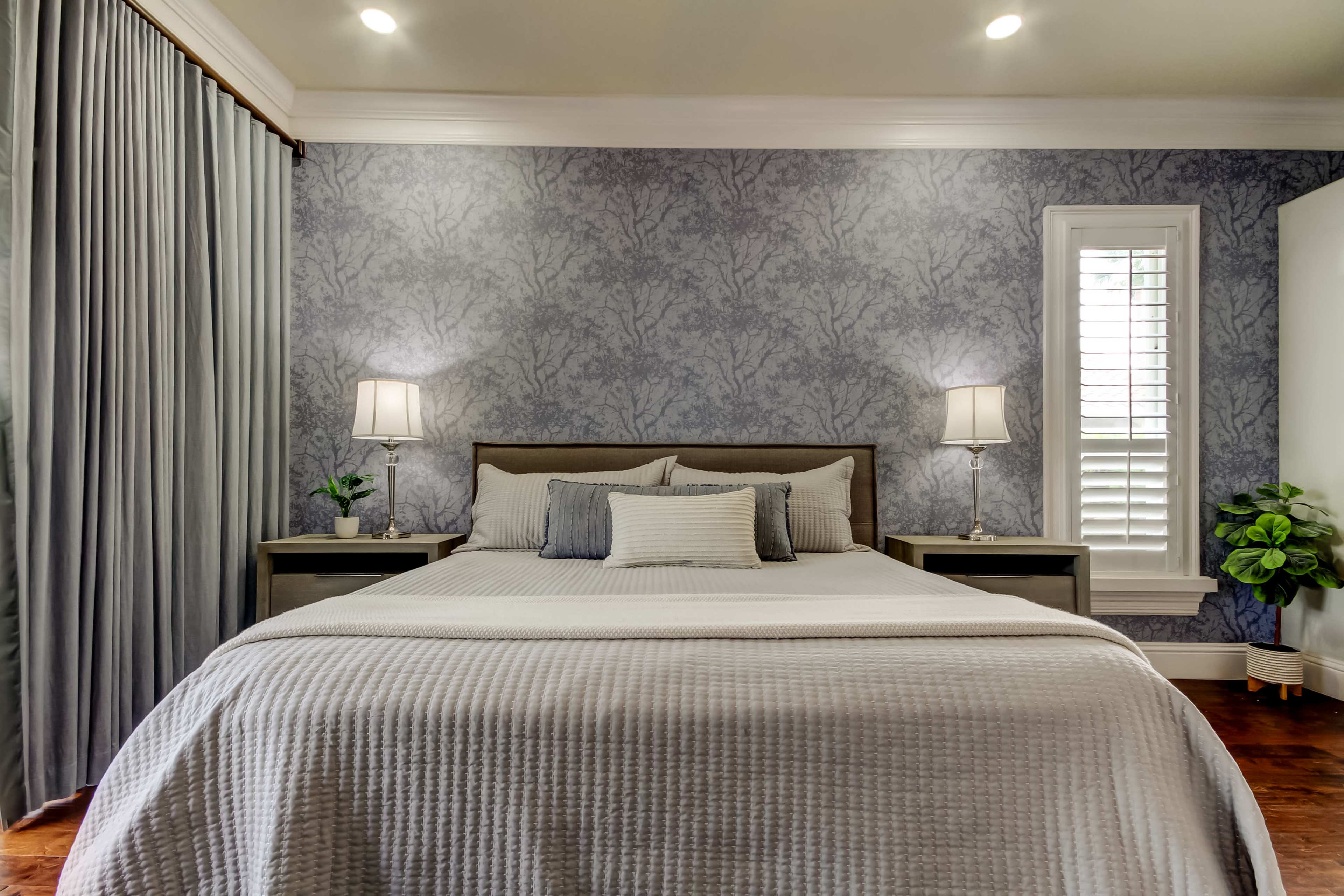 The image shows a neatly arranged bedroom with a bed, two bedside lamps, and a decorative plant, set against a patterned wall.
