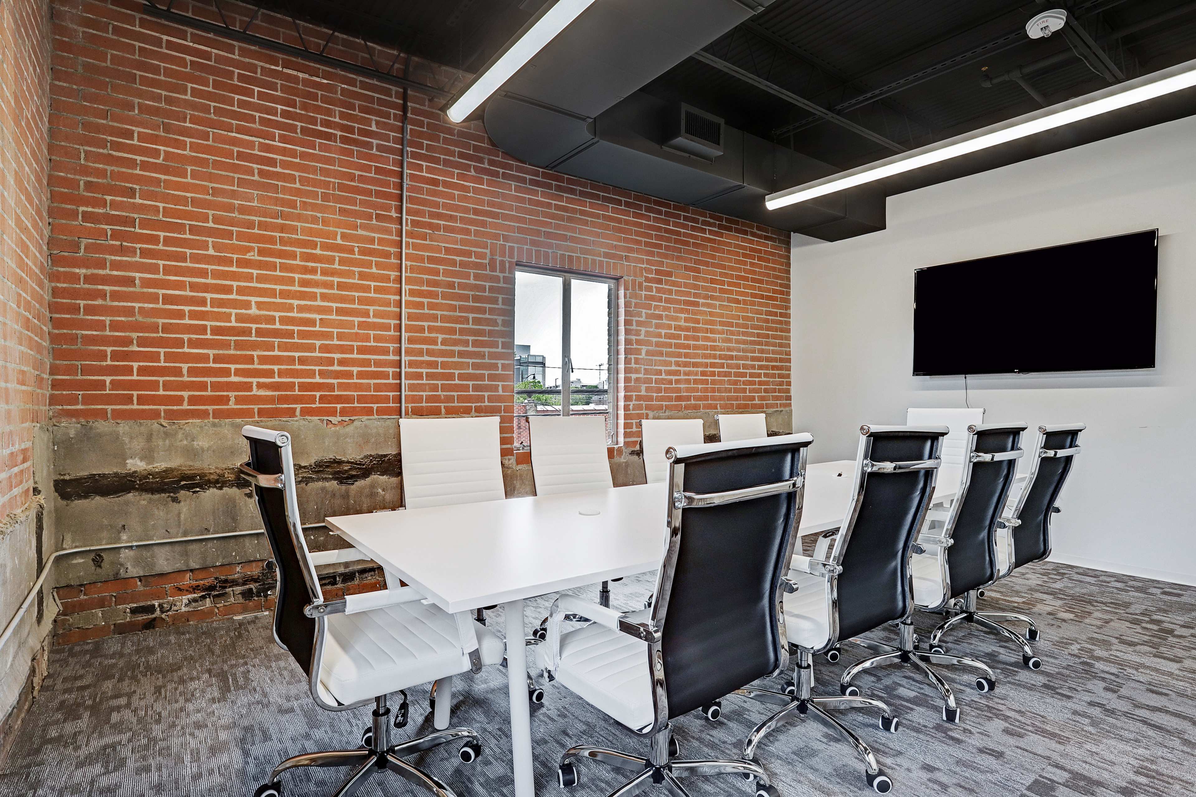 A modern conference room features a long white table surrounded by twelve black ergonomic chairs, with exposed brick walls and a large screen mounted on the opposite wall.