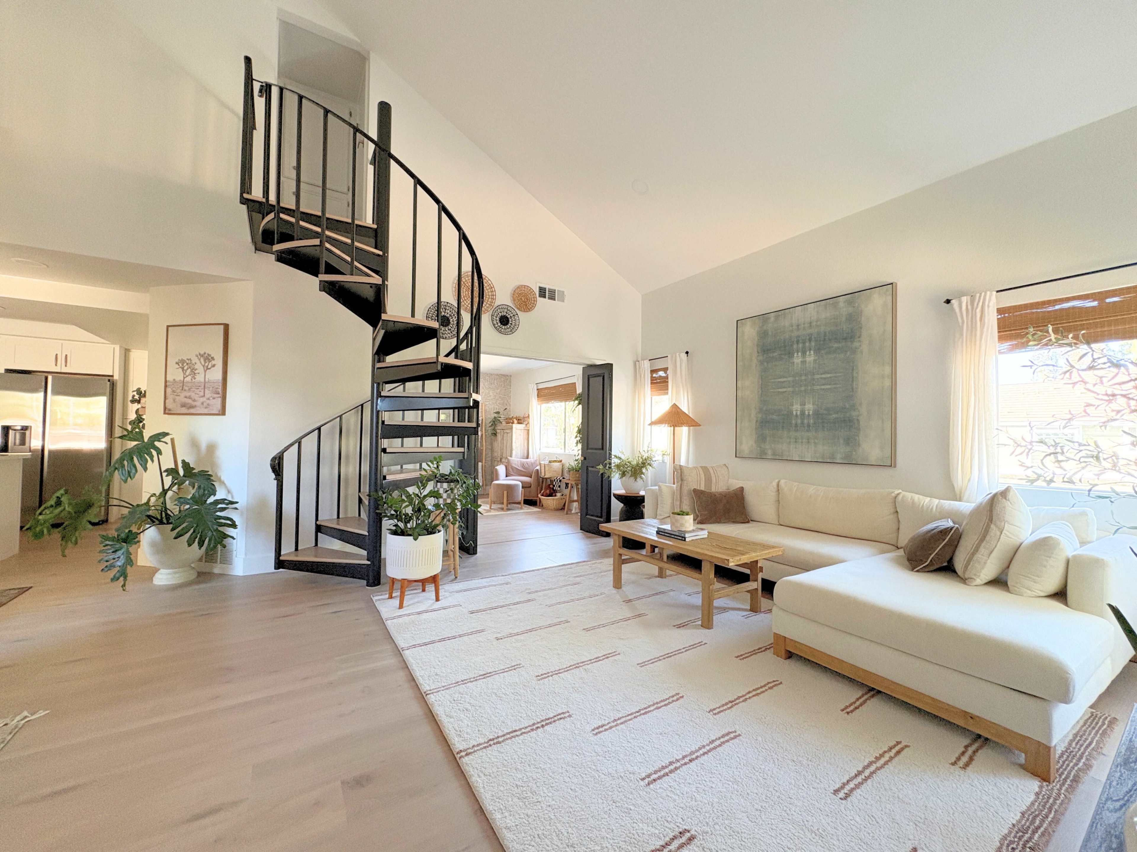 The image shows a bright and modern living room featuring a spiral staircase, a large sectional sofa, a coffee table, and various plants.