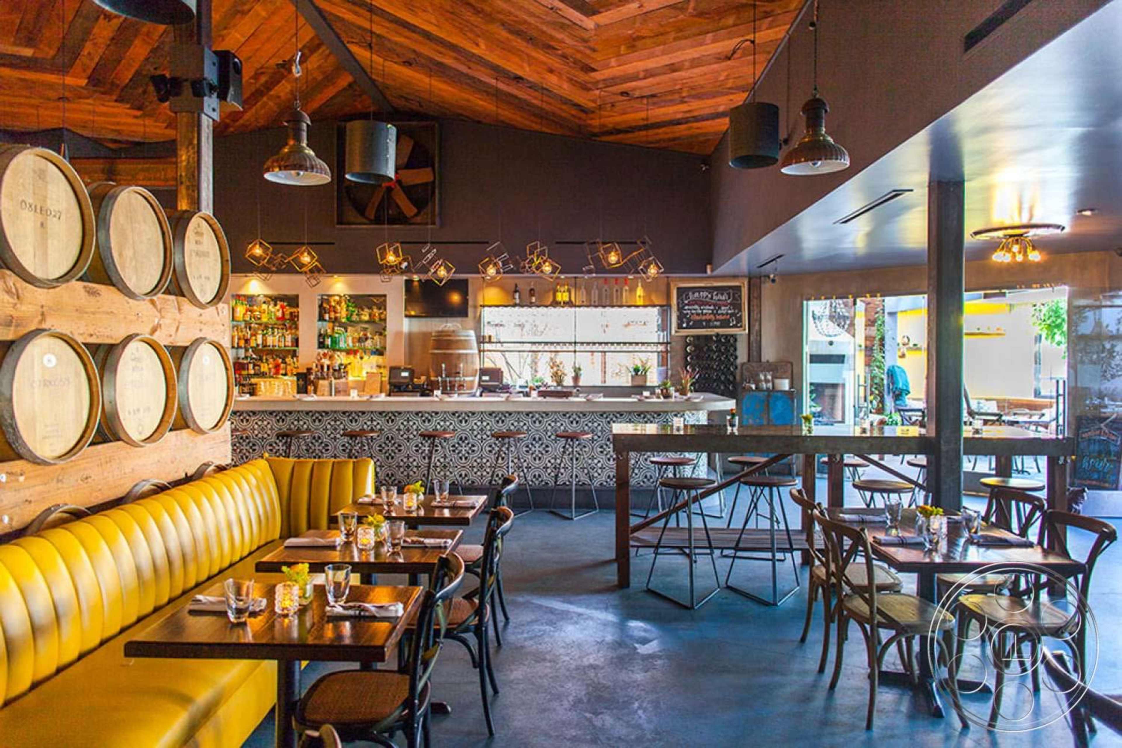 The image shows a modern restaurant interior with wooden accents, a bar, and tables arranged for dining.