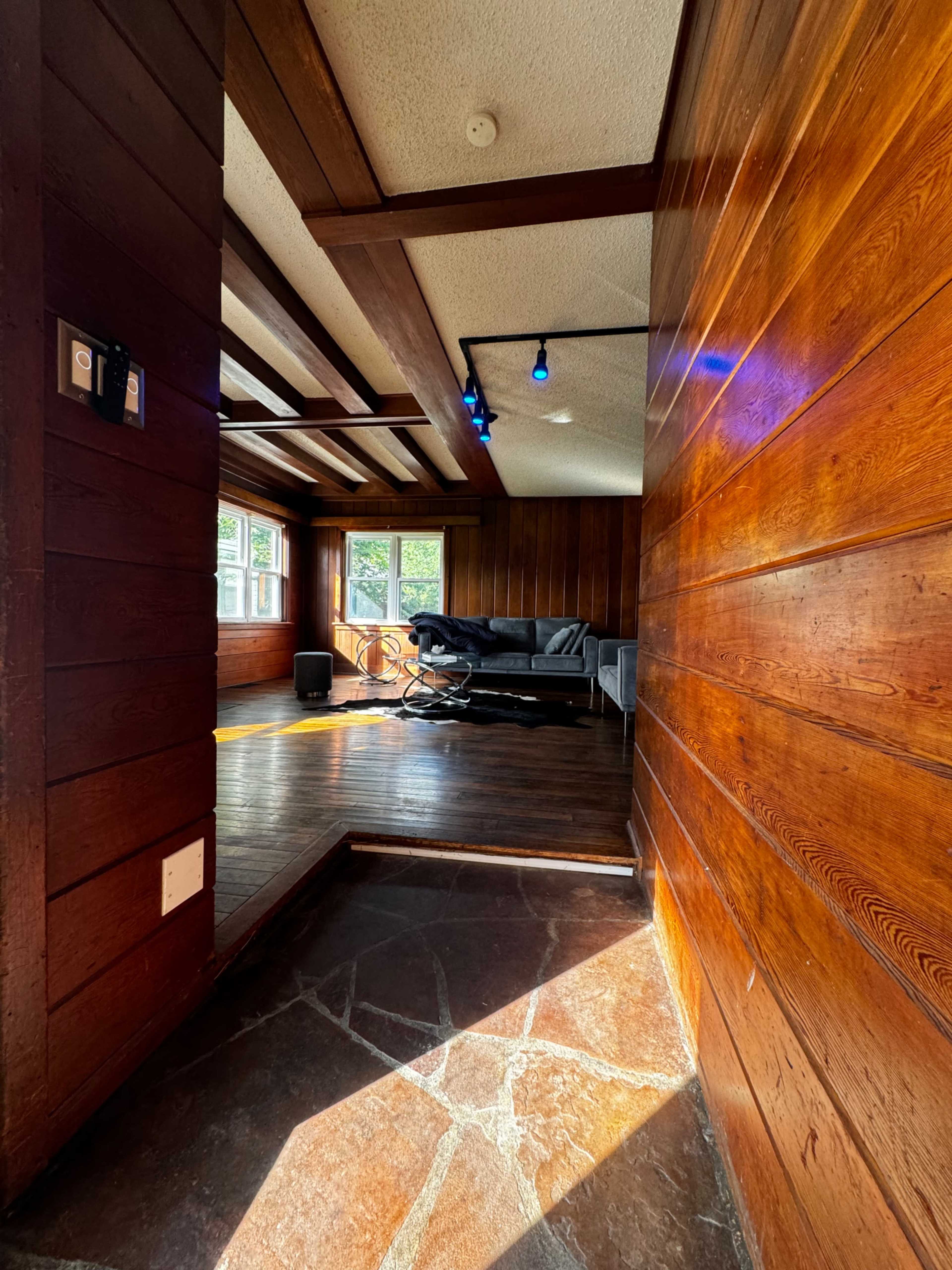 The image shows a wooden interior hallway leading into a well-lit living area with a dark couch and large windows.