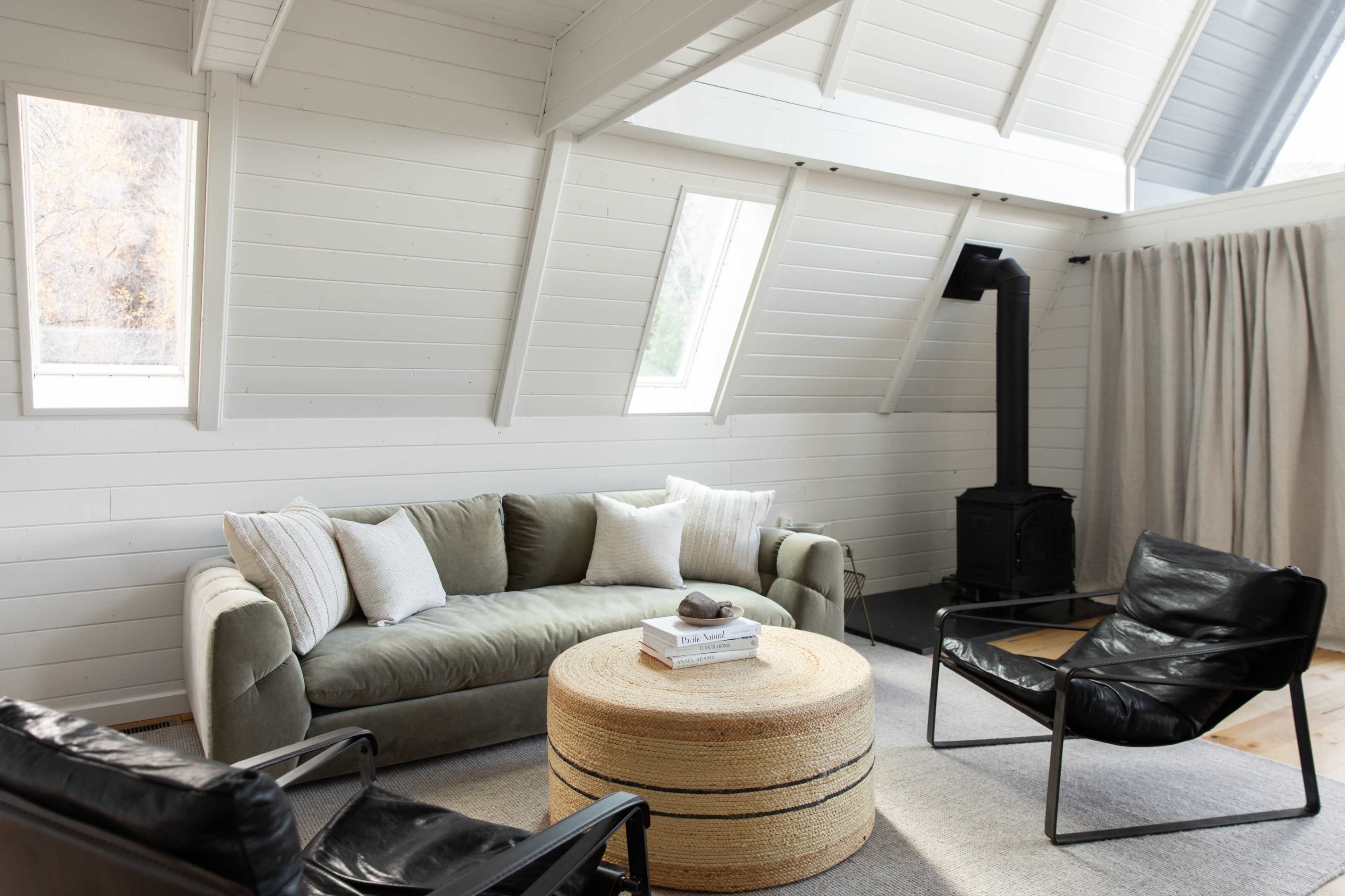 The room features a light-colored interior with an angular ceiling, a green sofa, two black chairs, and a round woven coffee table.