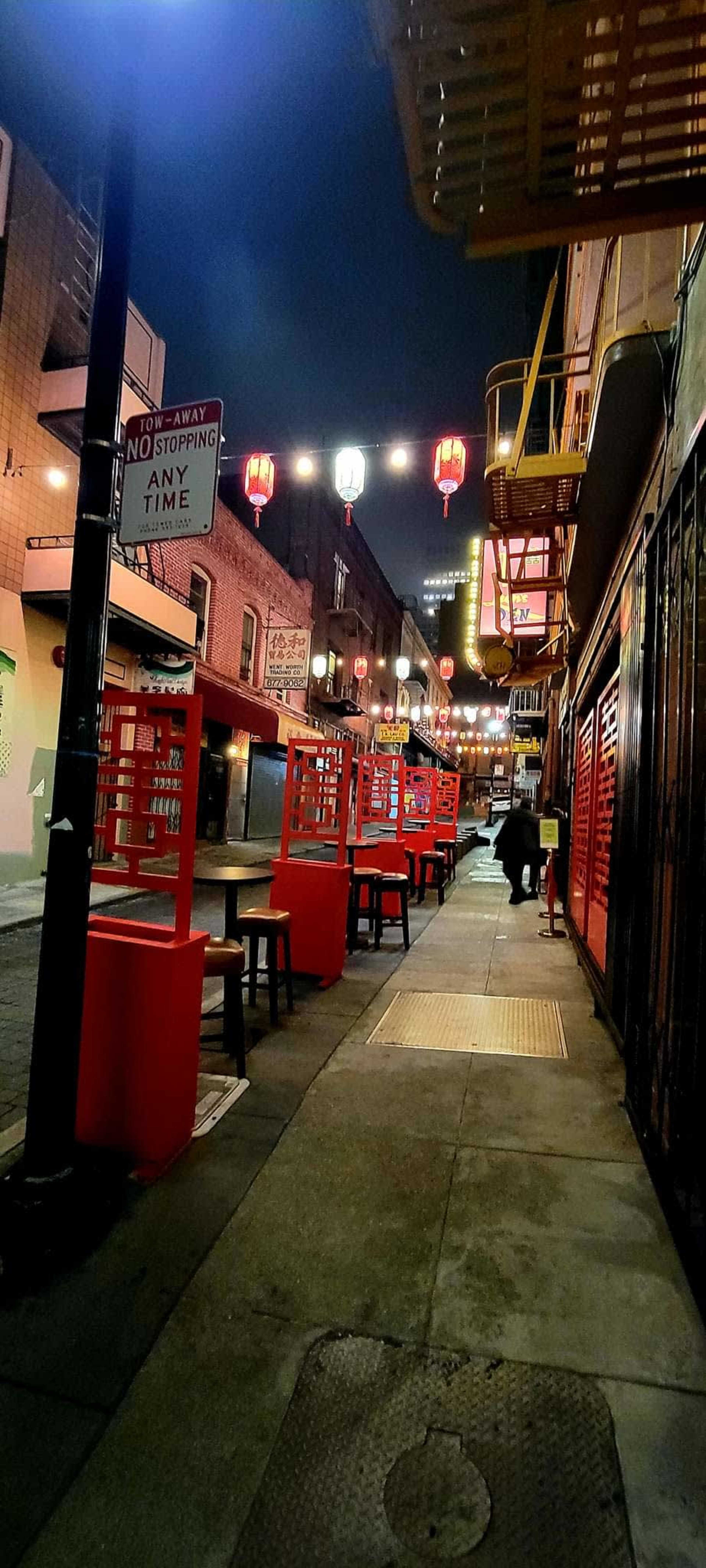 A quiet street in Chinatown features red tables and chairs lined along the sidewalk, illuminated by lanterns overhead.