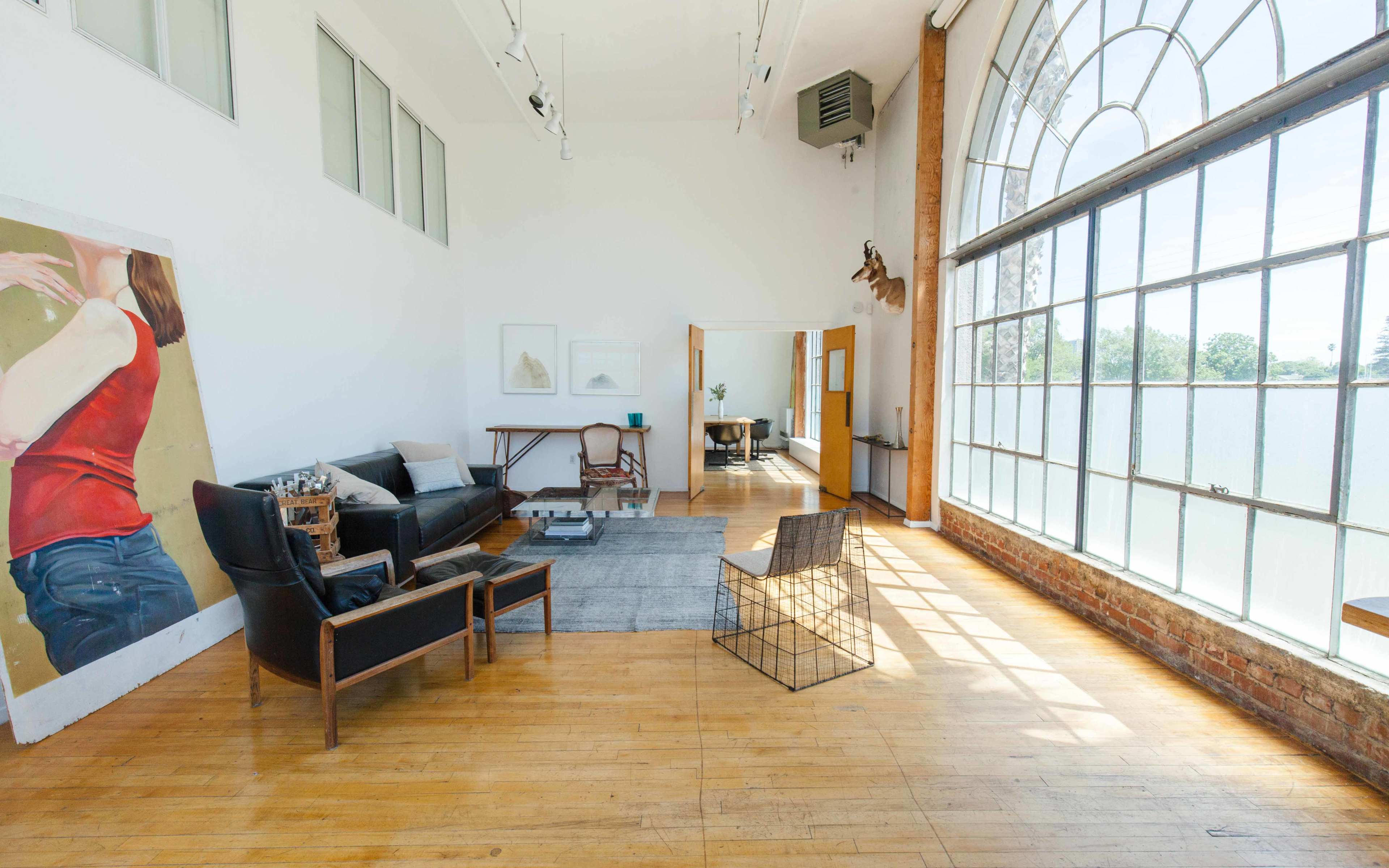 The image shows a spacious, brightly lit room with large windows, featuring a seating area with a sofa and two chairs, a painting on the wall, and a dining area in the background.
