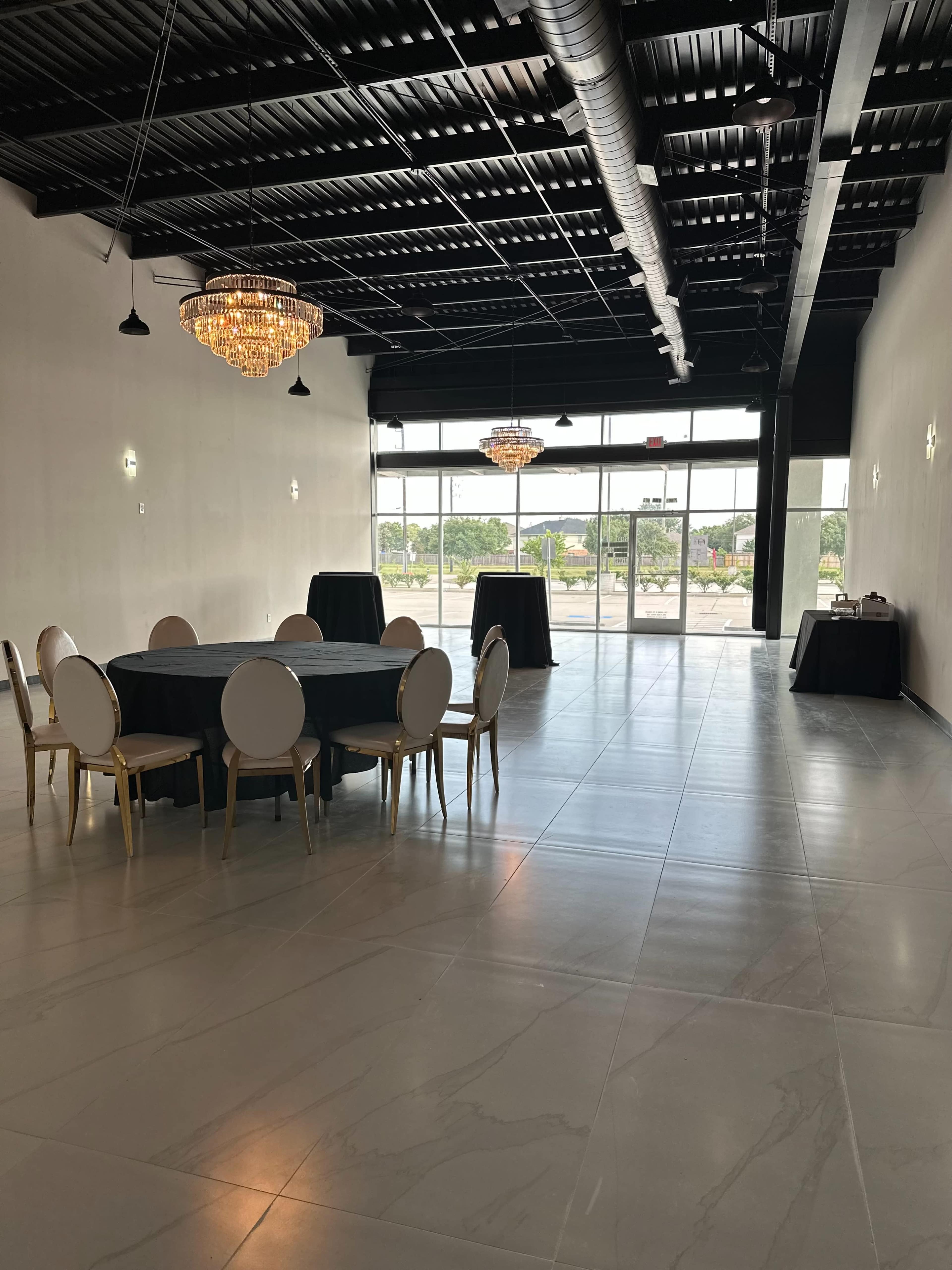 An empty event space with round tables, elegant chandeliers, and large windows allowing natural light to enter.