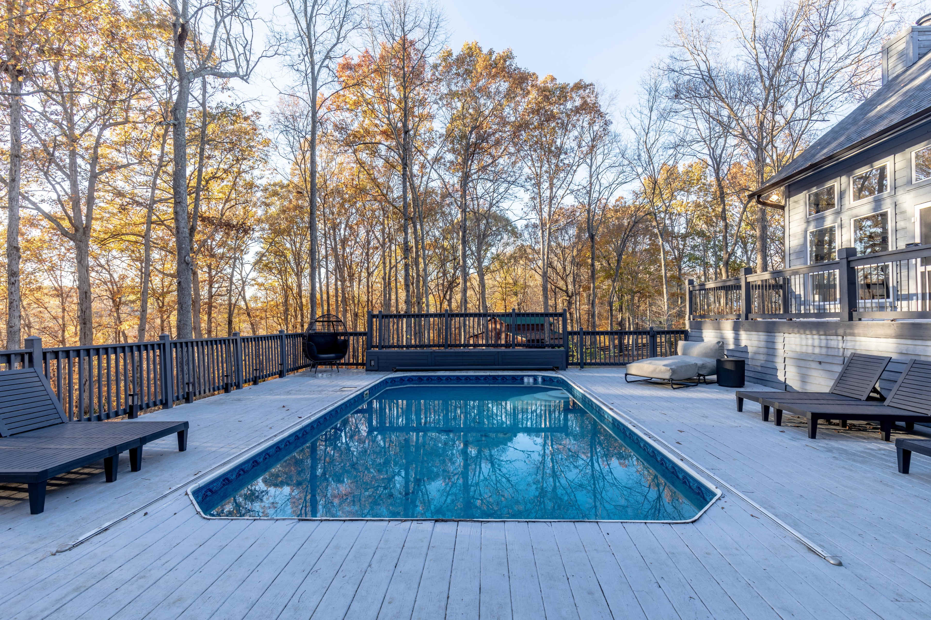 The image shows a rectangular swimming pool surrounded by wooden decking and lounge chairs, set against a backdrop of trees with autumn foliage.