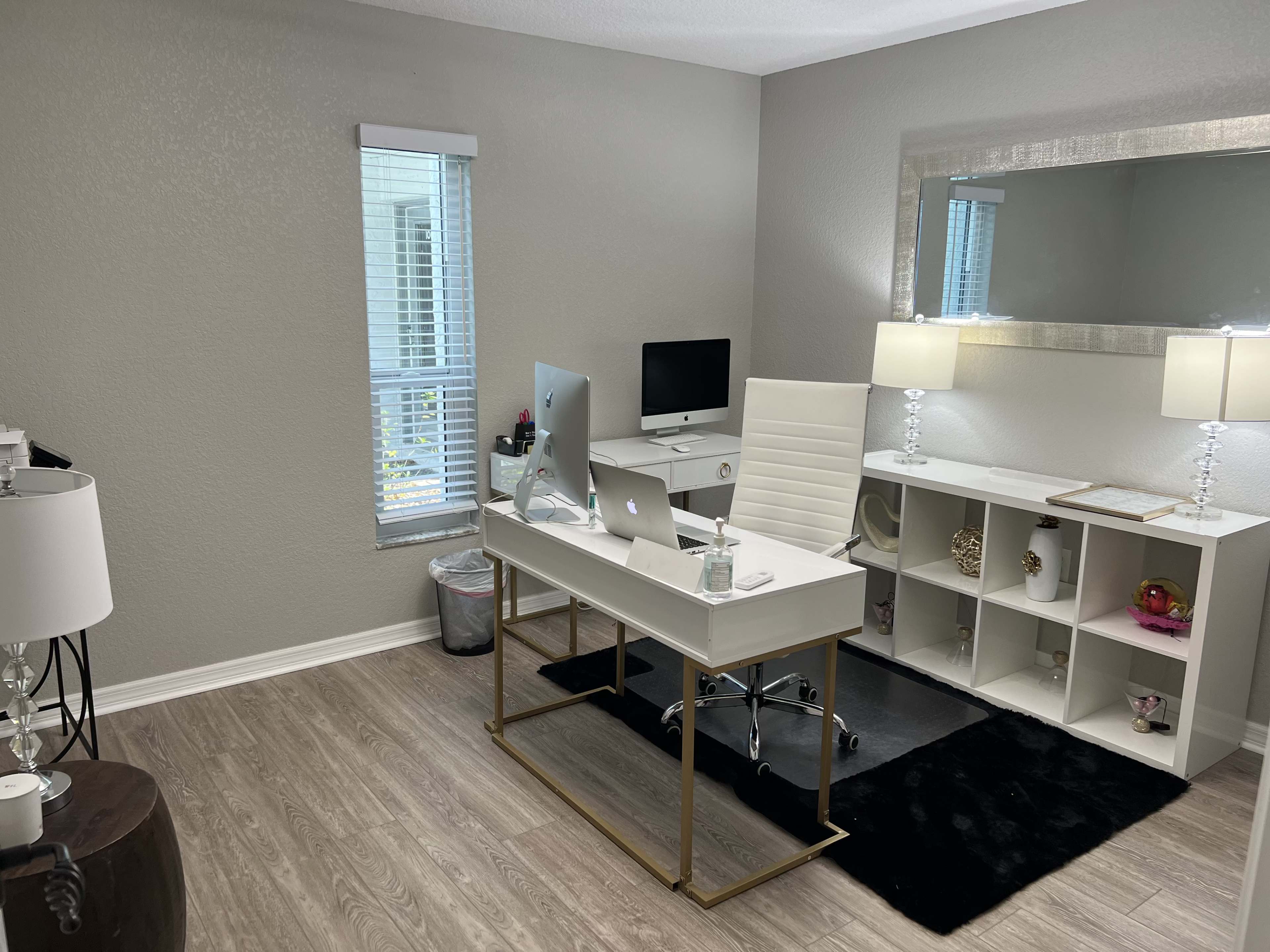 A modern office space features a white desk with a computer, a white ergonomic chair, a matching decorative table with lamps, and a storage unit against a light-colored wall.