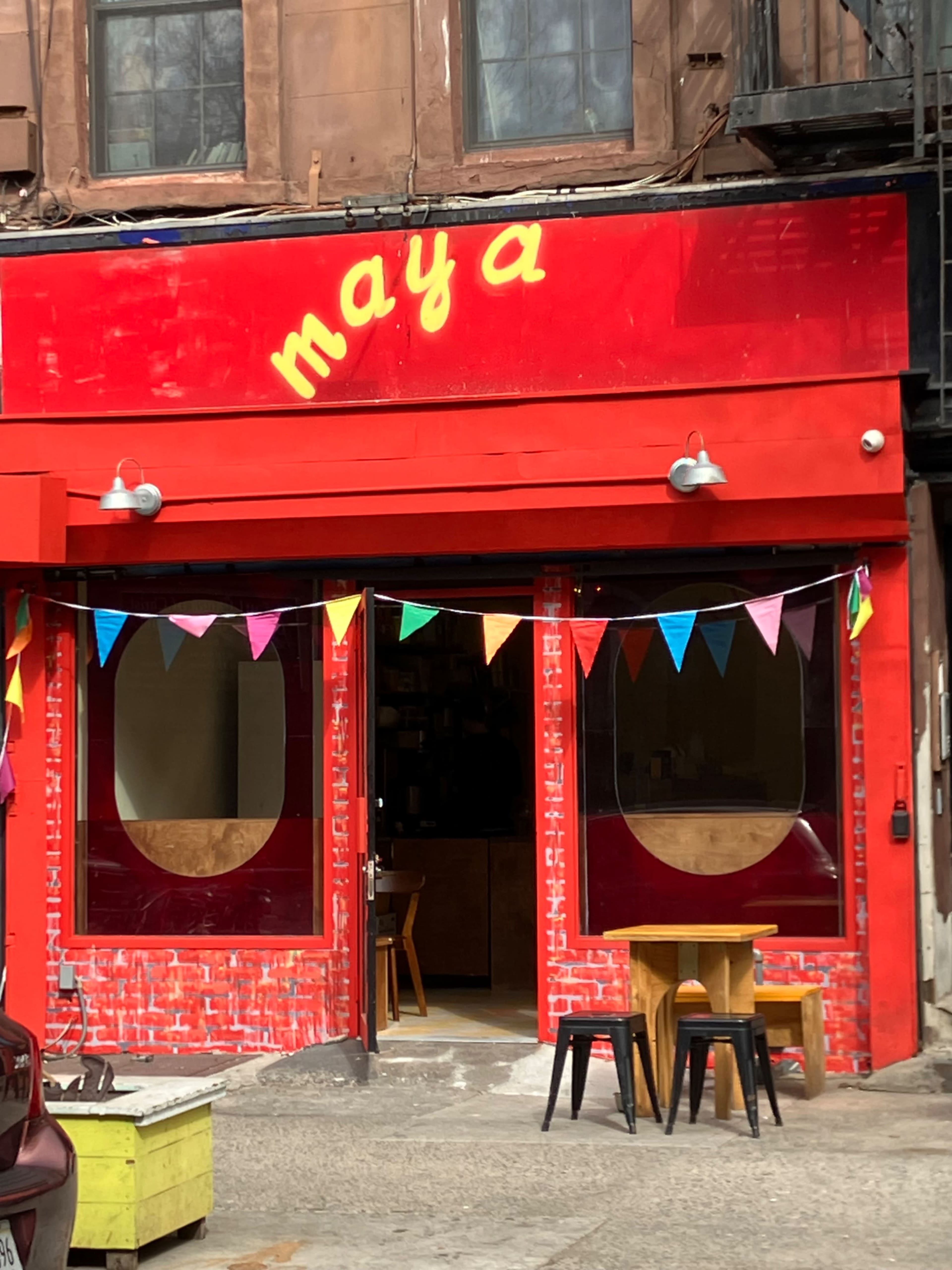 The image shows the entrance of a brightly colored café named "maya," featuring a red façade with decorative bunting and two small tables outside.