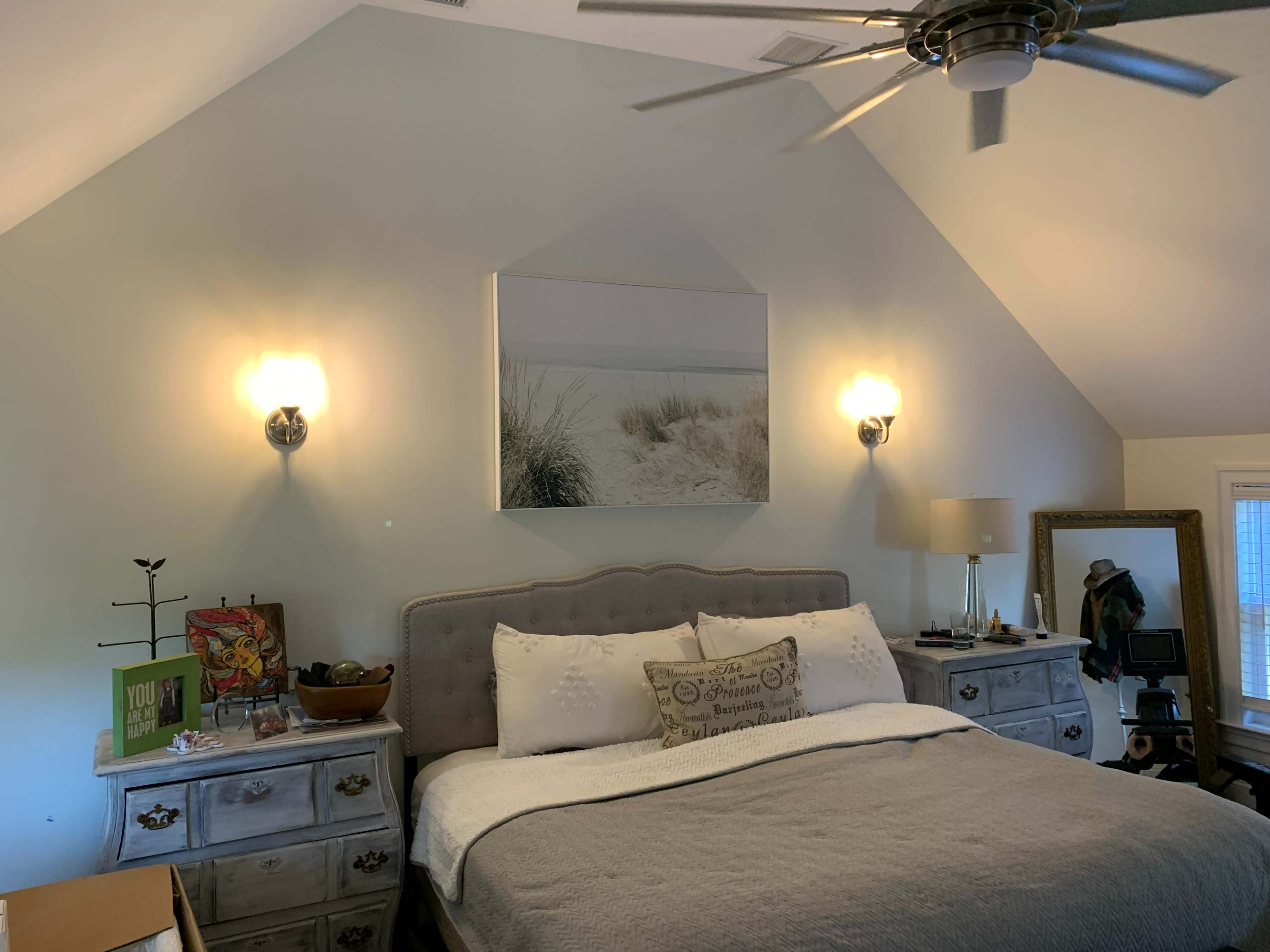 The image shows a well-furnished bedroom with a large bed, artwork on the wall, and light fixtures on either side of the bed.