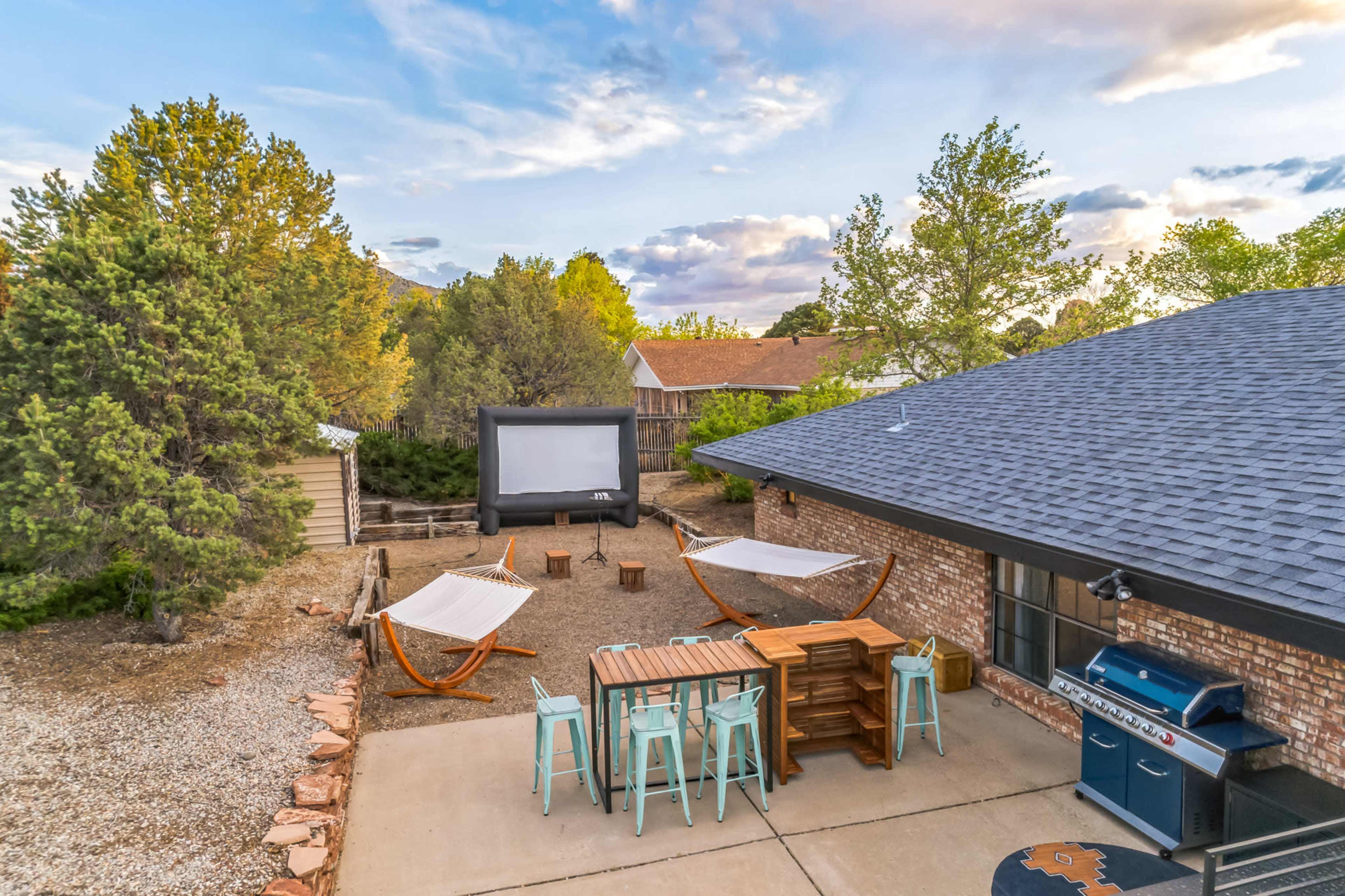 A backyard setup with hammocks, a large outdoor movie screen, seating, and a grill, surrounded by trees and a gravel area.