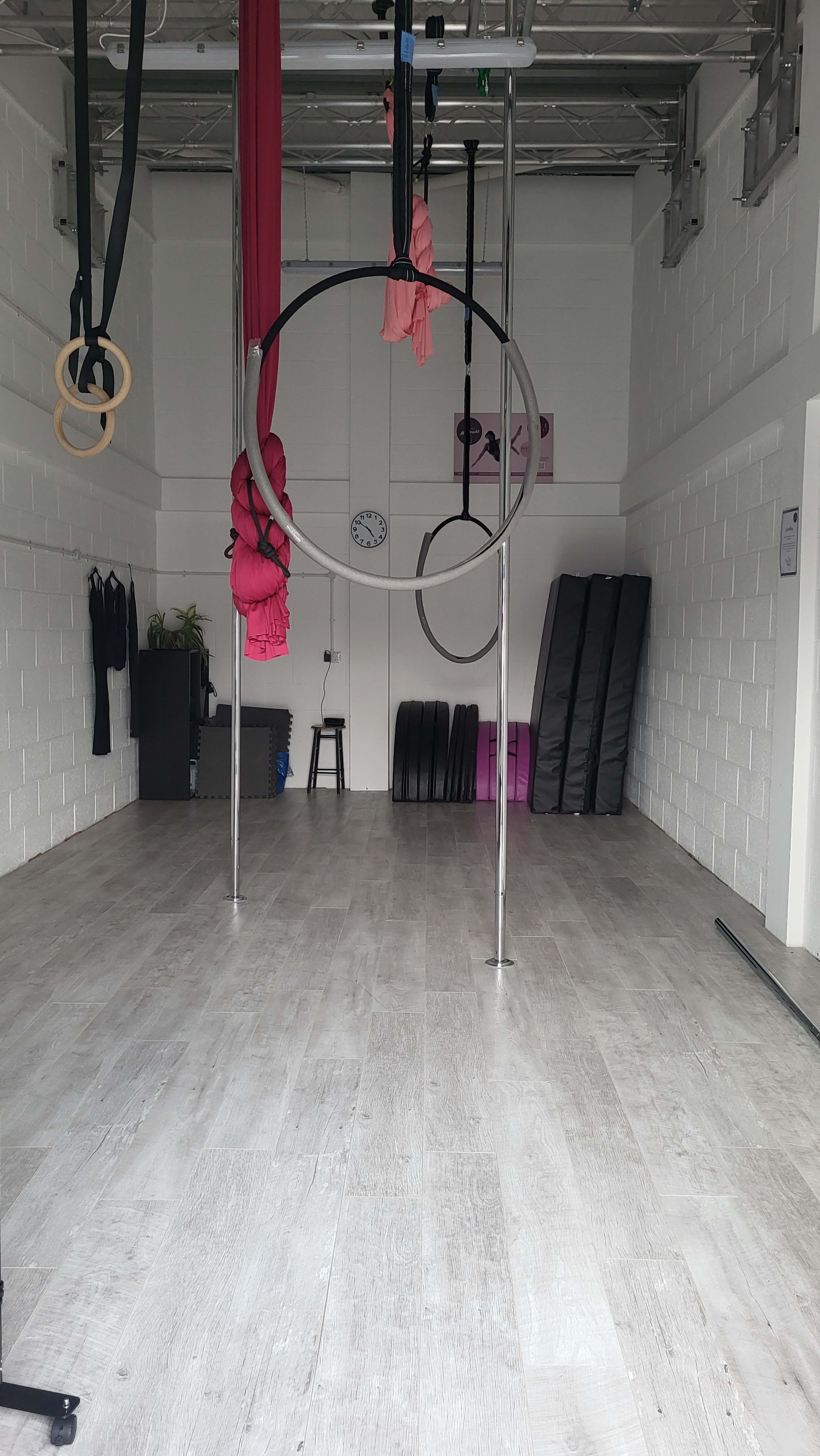 The image shows a spacious gym interior with aerial silks and rings suspended from a ceiling, along with exercise mats stacked against the wall.