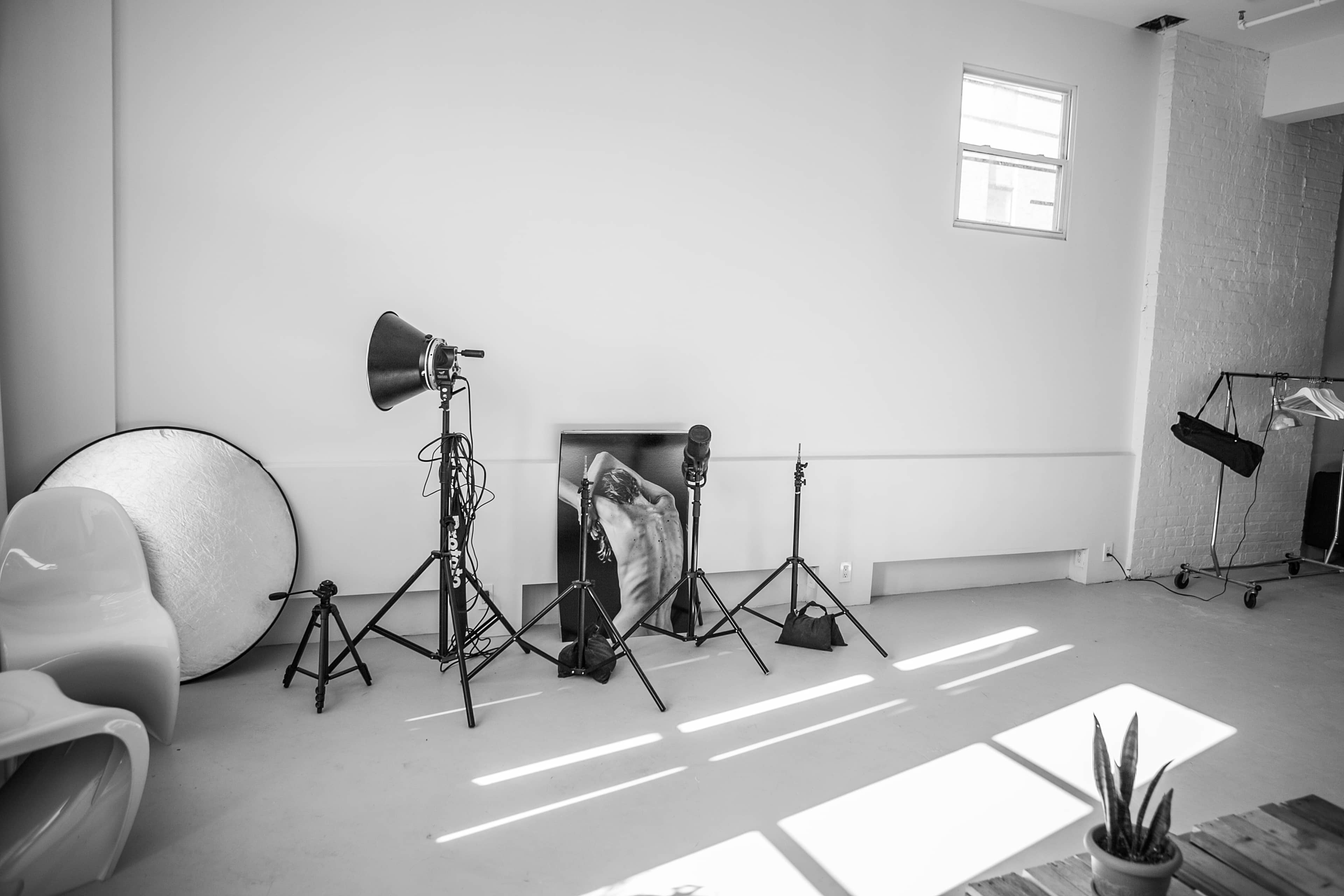 The image shows a minimalist photo studio with lighting equipment and a framed black-and-white photograph on the wall.