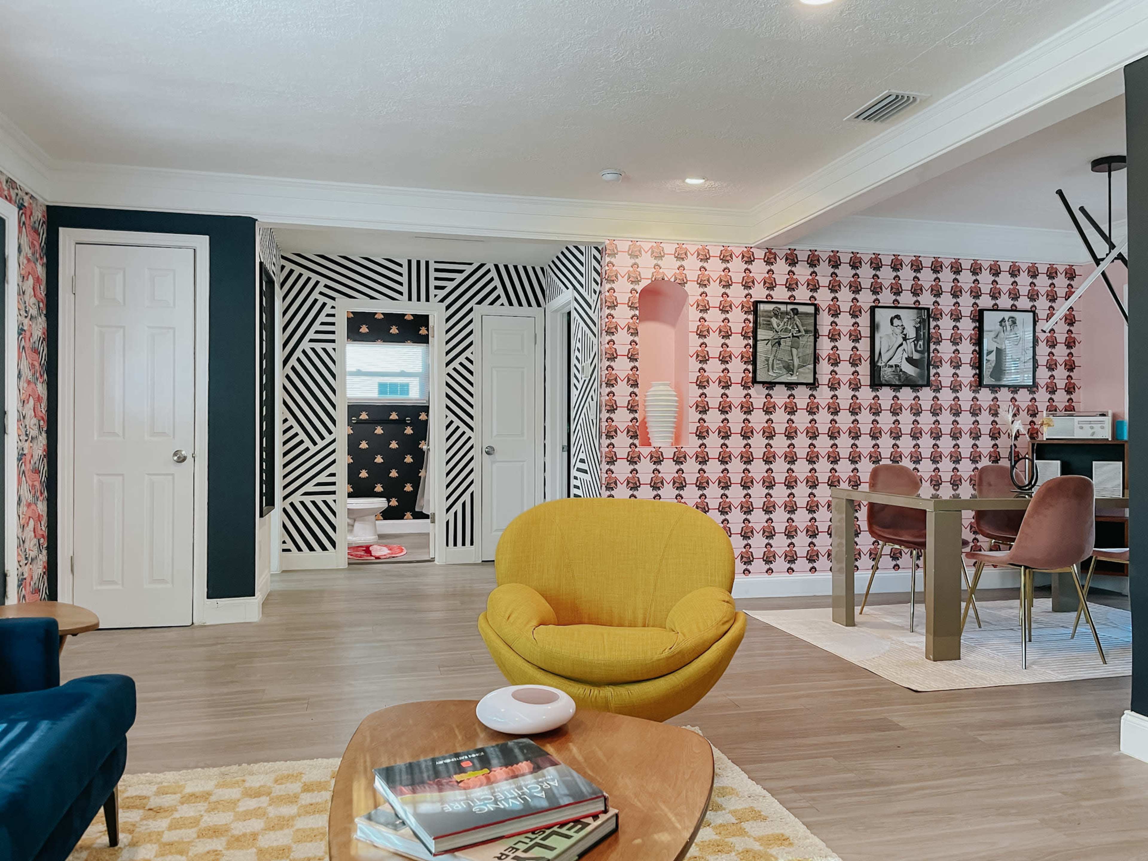 The image shows a brightly decorated living space featuring colorful wallpaper, a yellow armchair, a wooden coffee table stacked with books, and a dining area with a dark table against contrasting wall patterns.