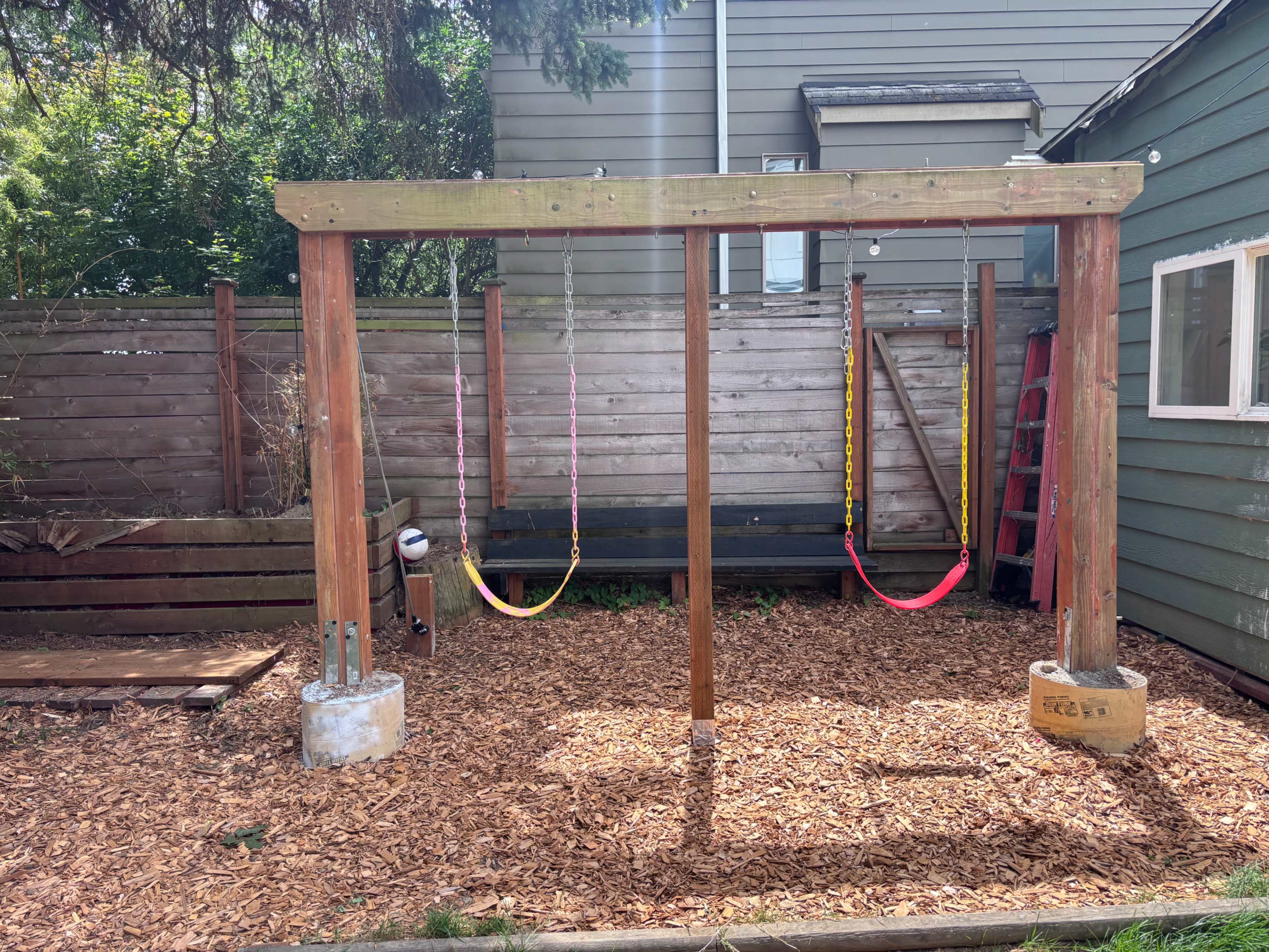 The image shows a wooden swing set with two swings, set on a bed of wood chips in a backyard surrounded by a wooden fence and a house in the background.