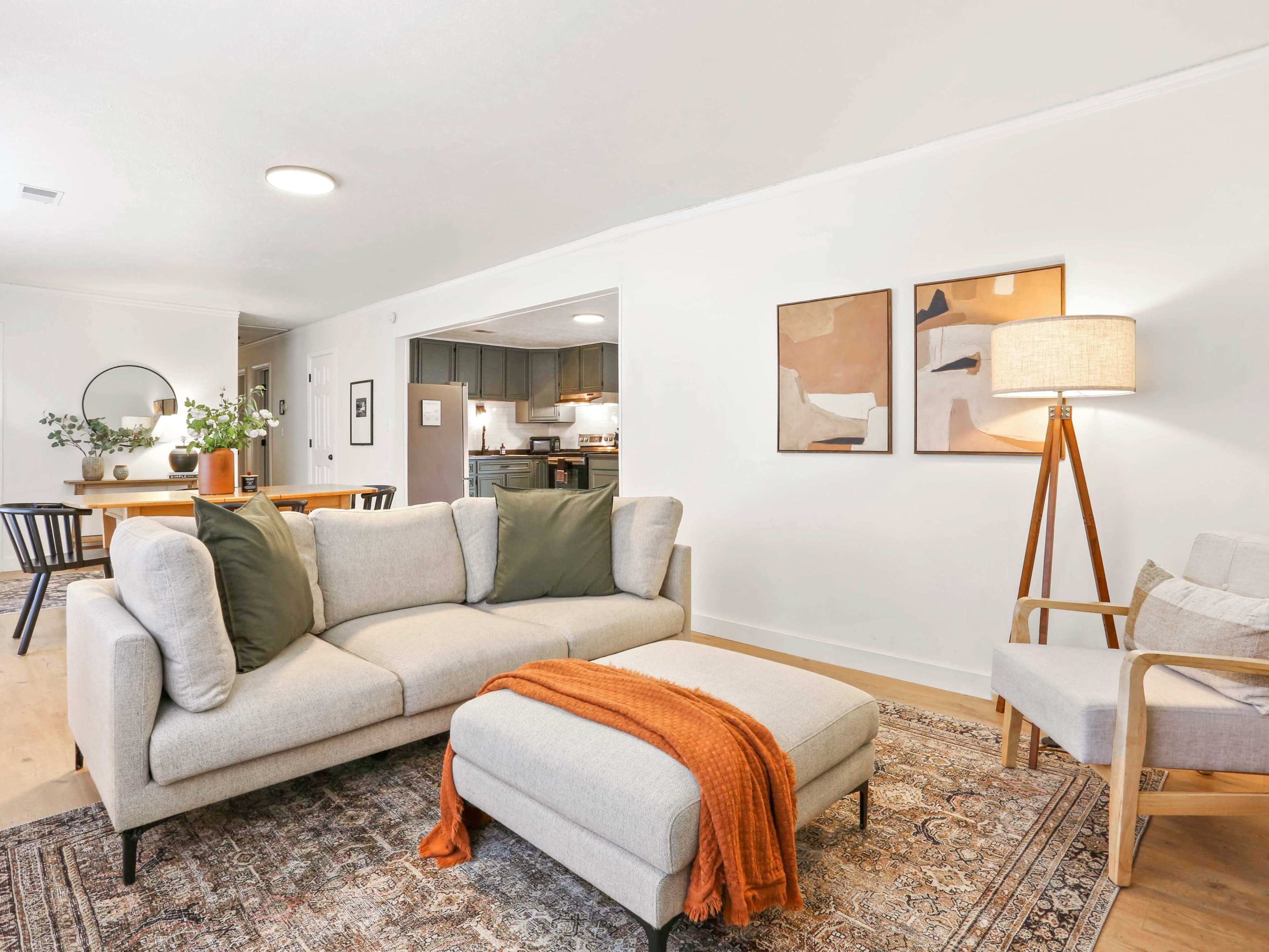 A modern living room features a light gray sectional sofa with green cushions, an orange throw, and a floor lamp, alongside a dining area visible in the background.