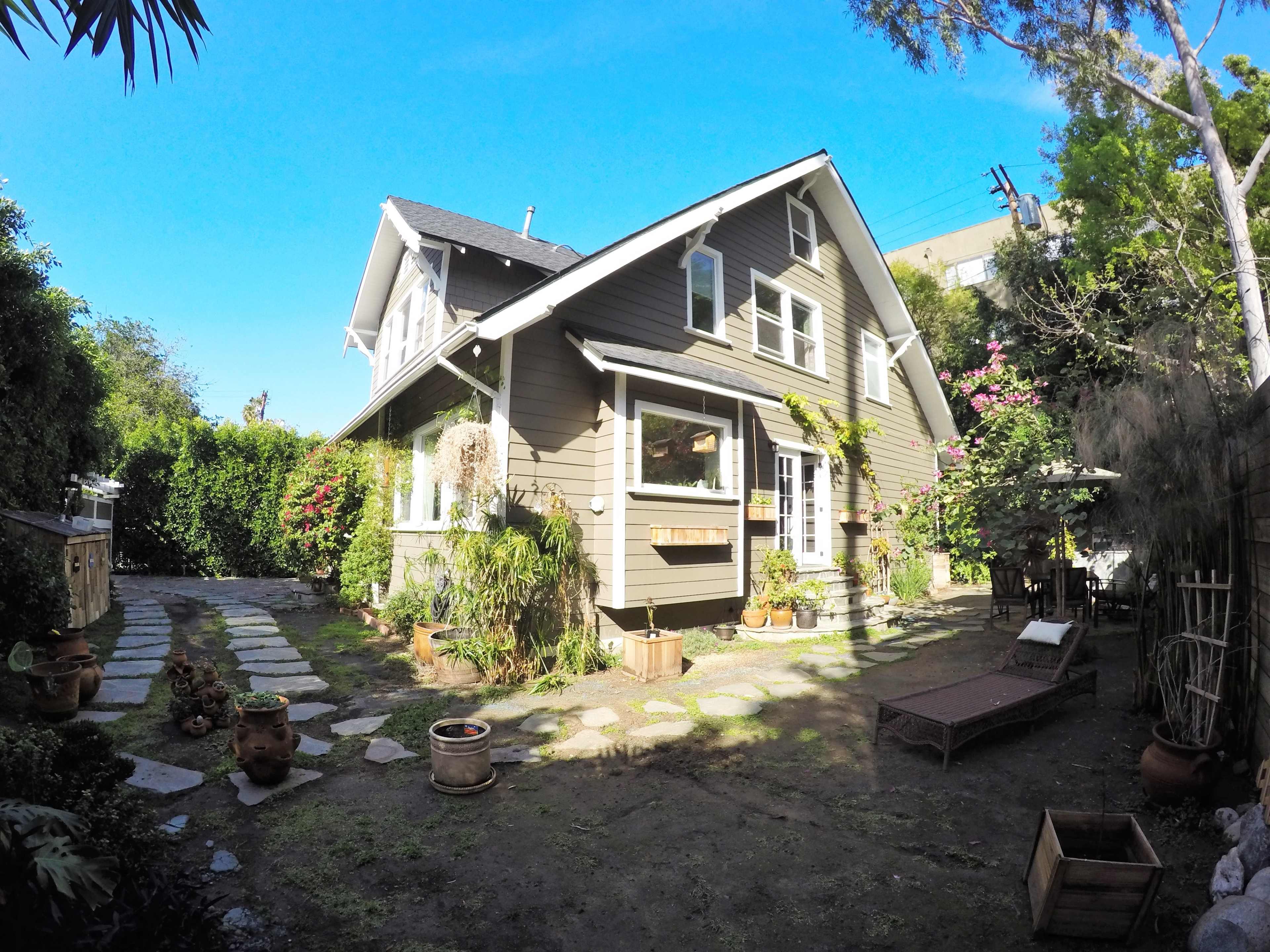 A two-story wooden house with a sloping roof is surrounded by a garden featuring stone pathways, potted plants, and a small seating area.