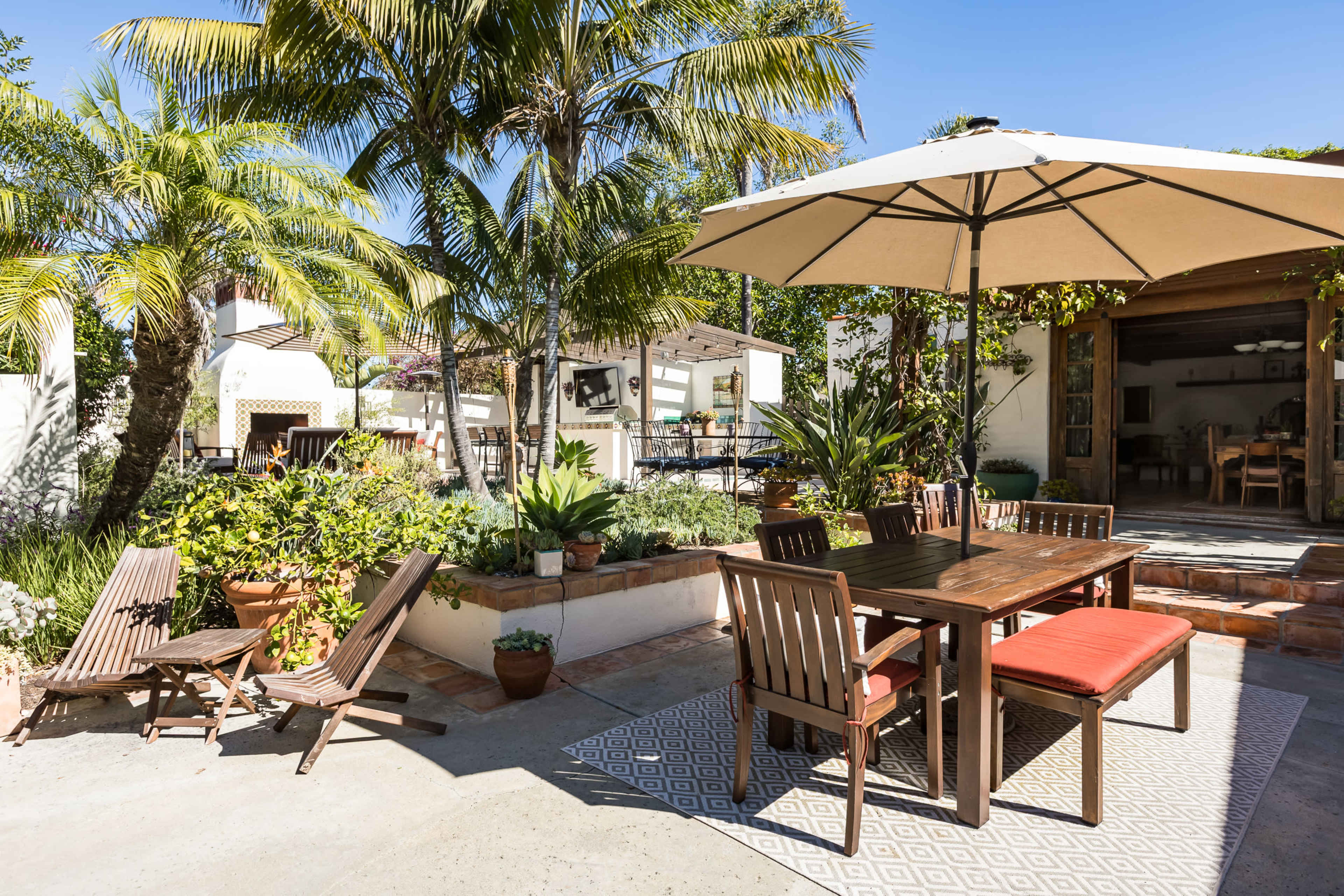 A sunlit outdoor patio features a wooden table with chairs, an umbrella, potted plants, and two lounge chairs set against a backdrop of palm trees and a house.