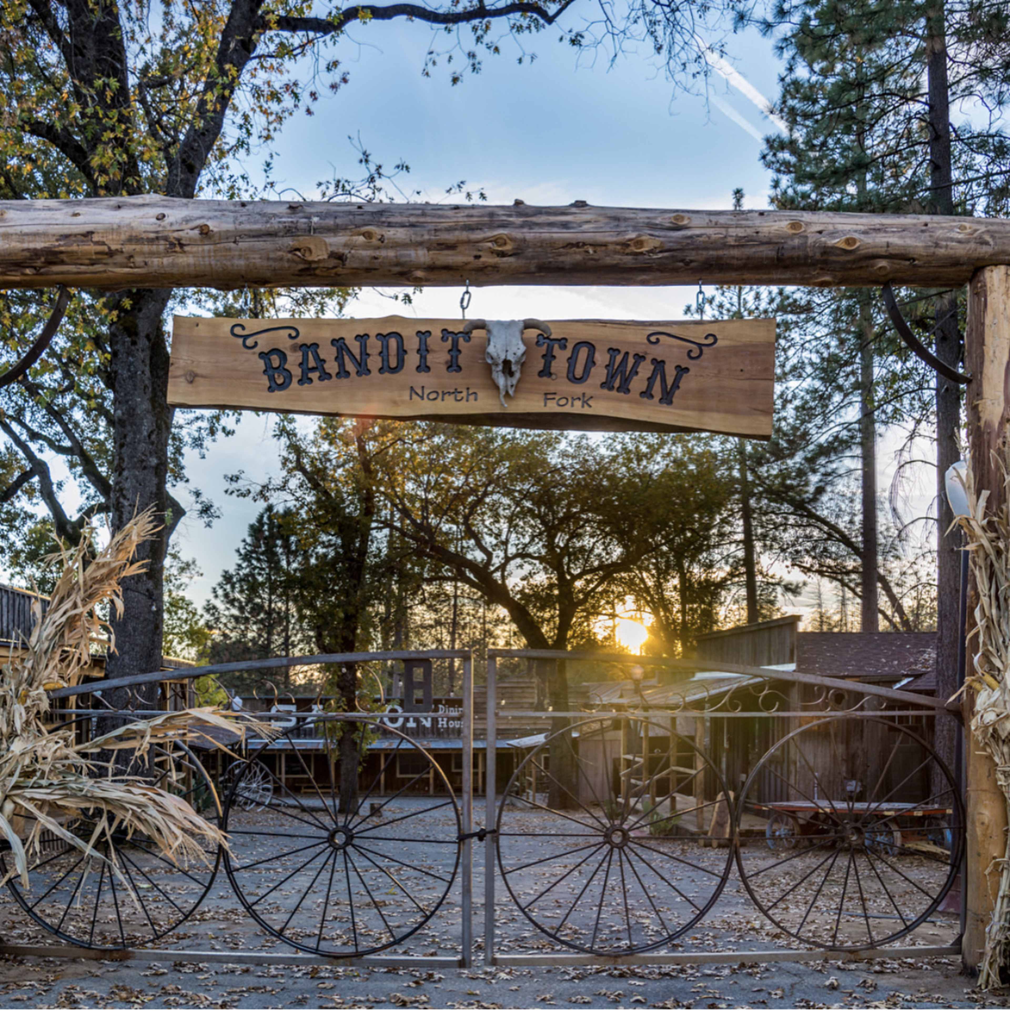 The entrance to Bandit Town in North Fork is marked by a wooden sign hanging from a rustic archway, set against a backdrop of trees and a sunset.