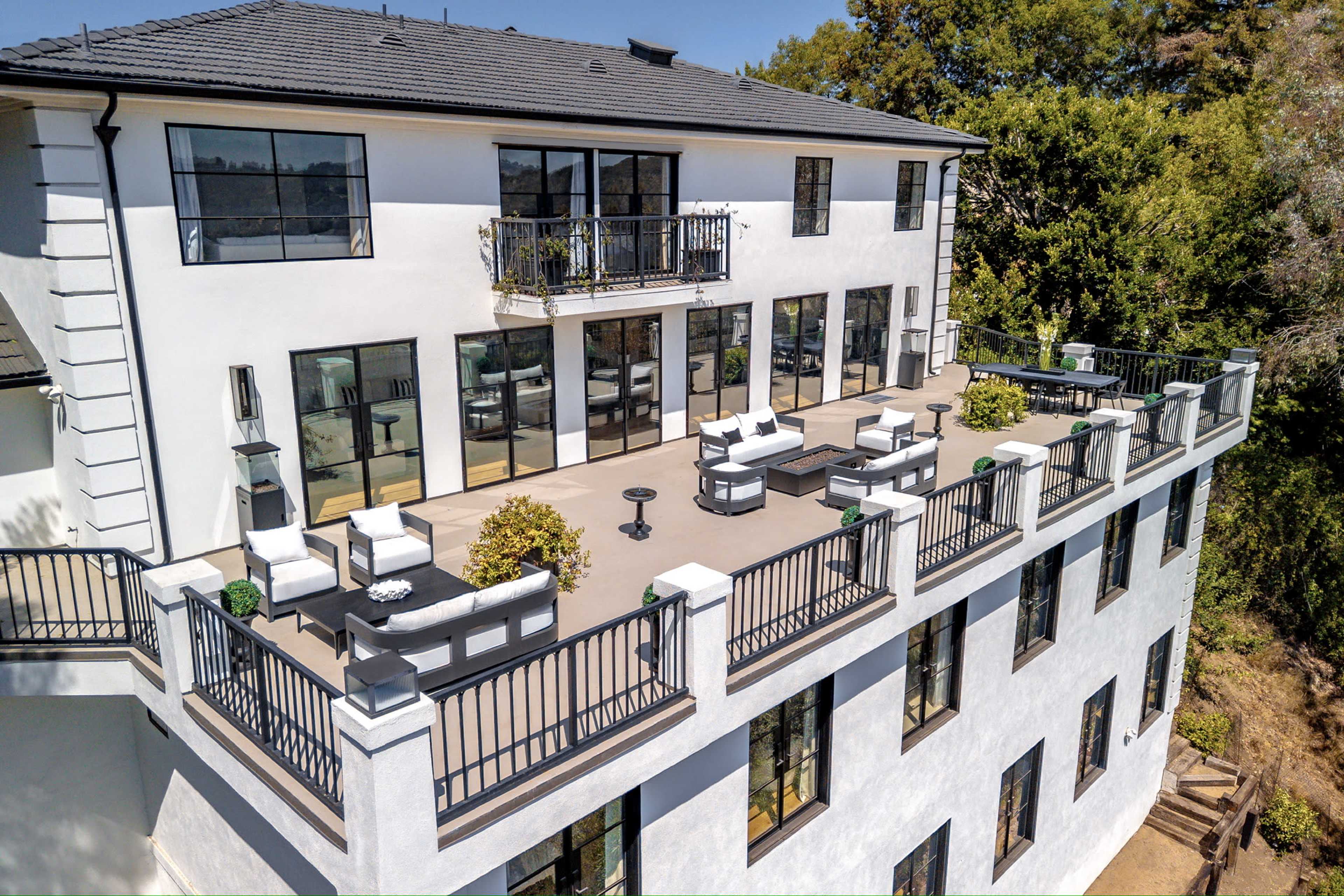 The image shows a large, modern white house with multiple balconies and a spacious terrace furnished with outdoor seating.