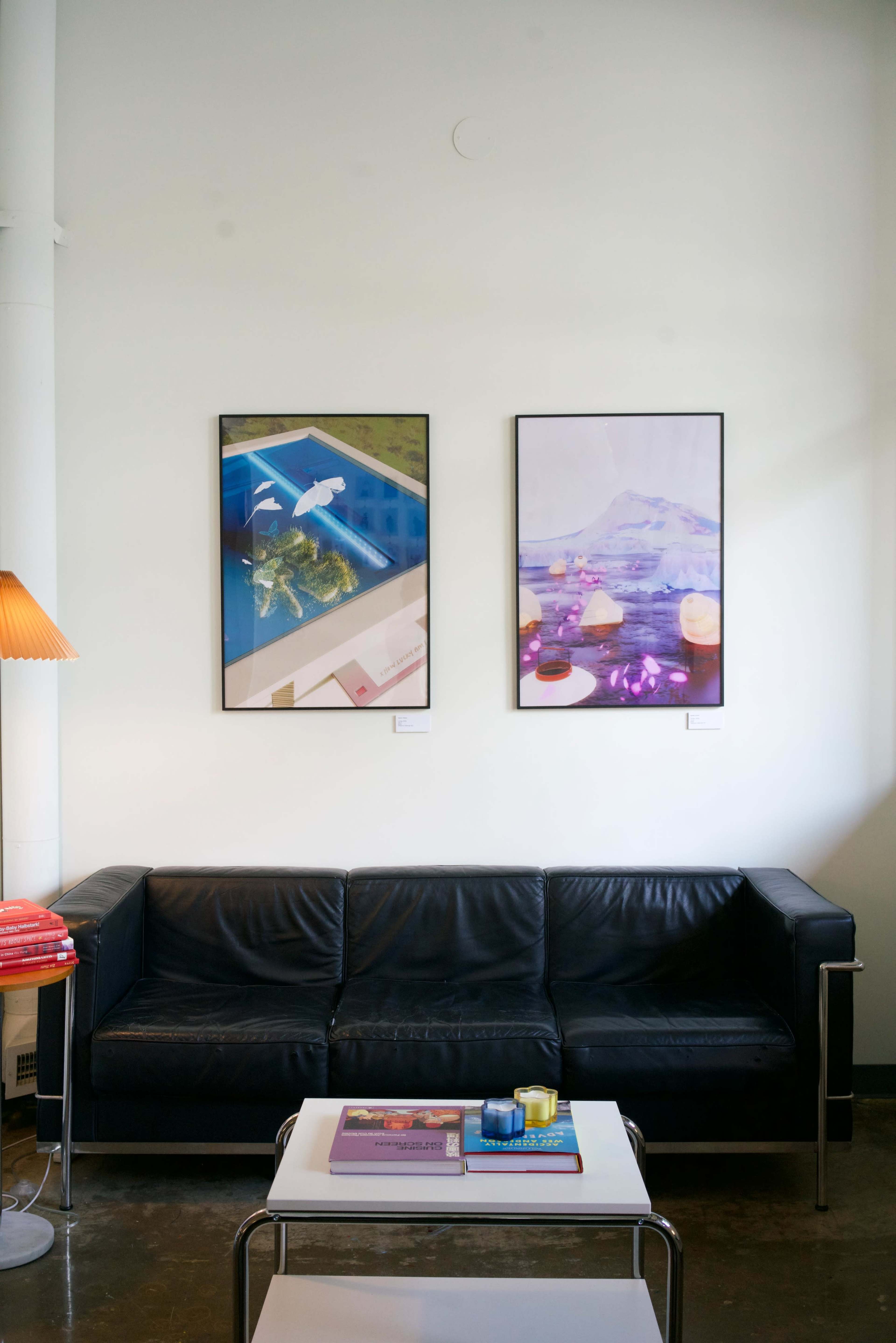 The image shows a minimalist living space featuring a black leather sofa, a coffee table with books, and two colorful framed artworks on the wall above.