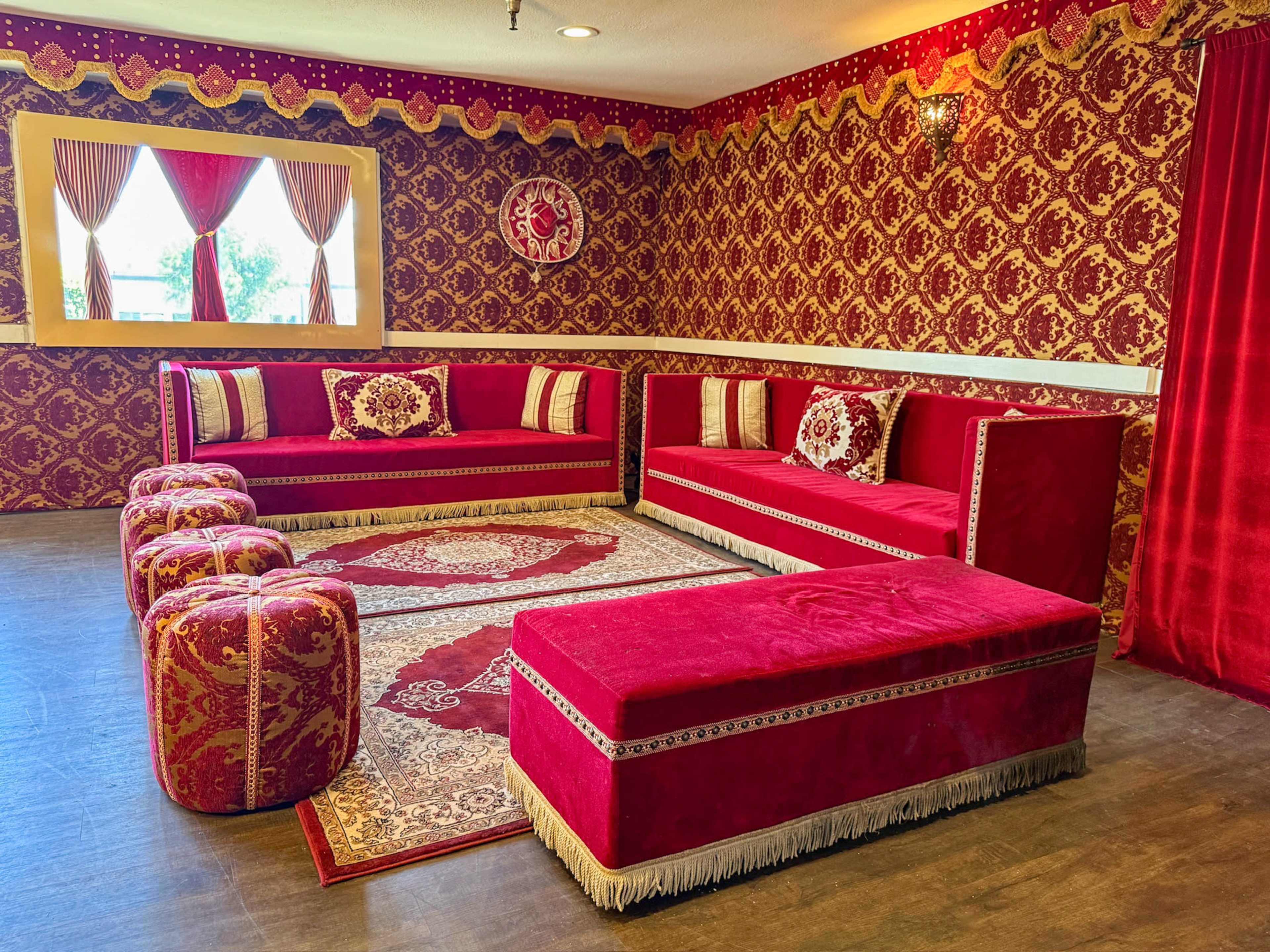 The image shows a room decorated with red and gold patterned wallpaper, featuring red velvet furniture, including a sofa, ottomans, and a large area rug.