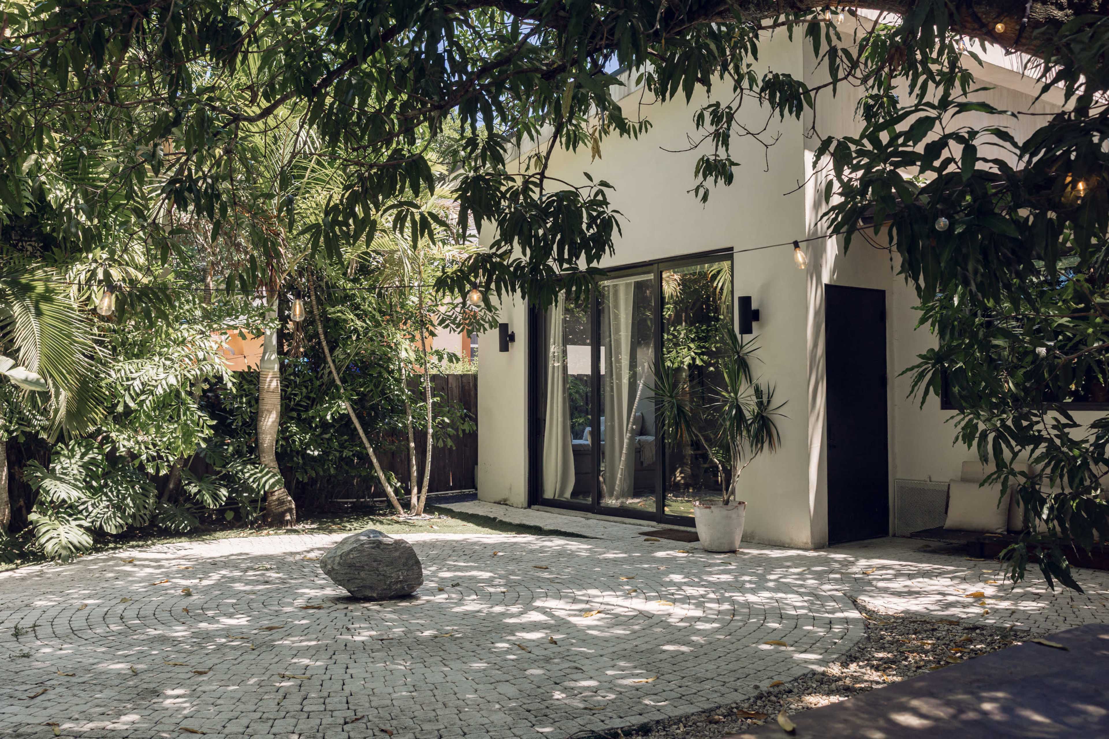 A modern house with large glass doors is surrounded by lush greenery and features a circular stone pathway with a boulder in the center.