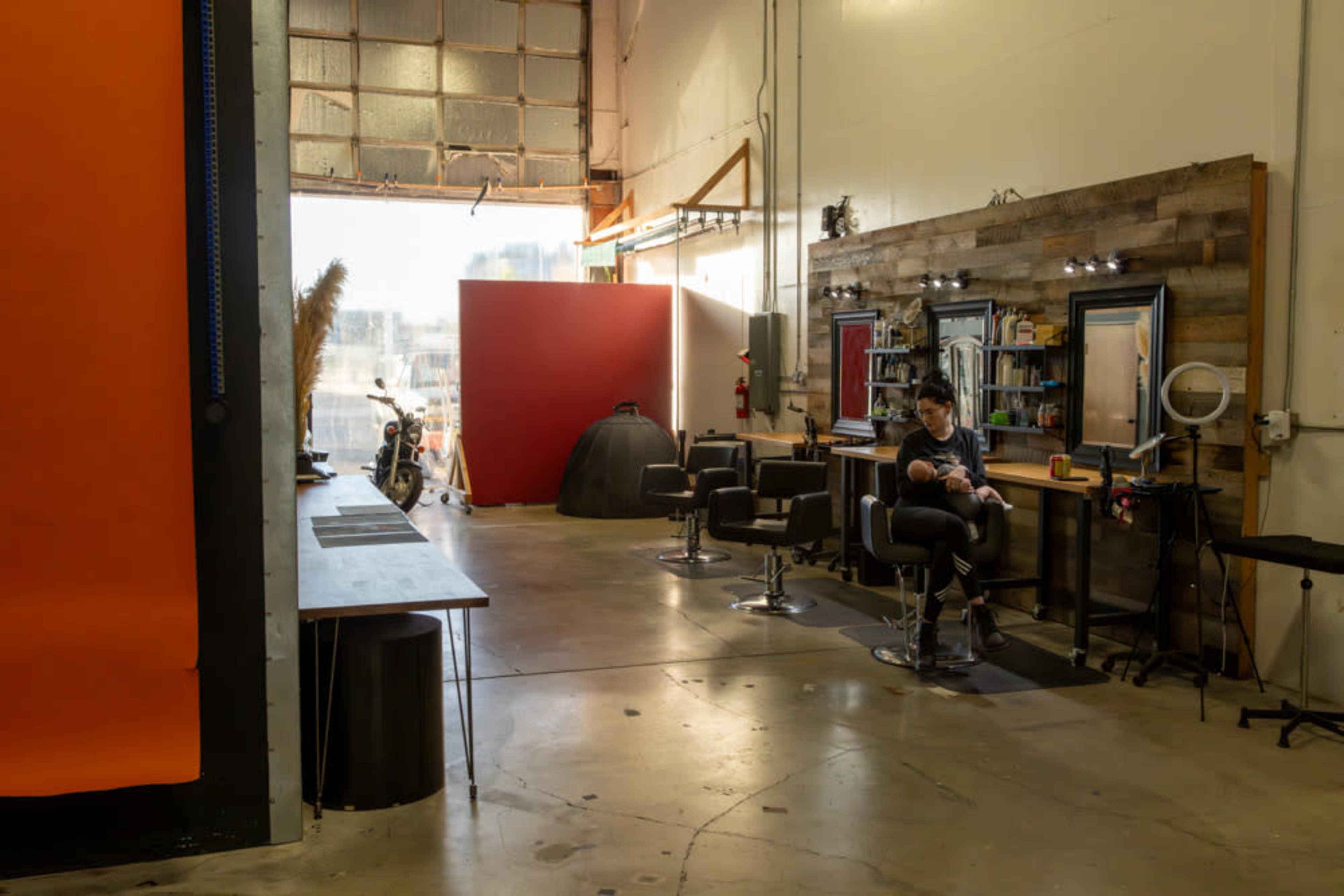 The image shows a minimalist hair salon with a row of black barber chairs, mirrors, and a wooden accent wall, bathed in natural light from an open garage door.