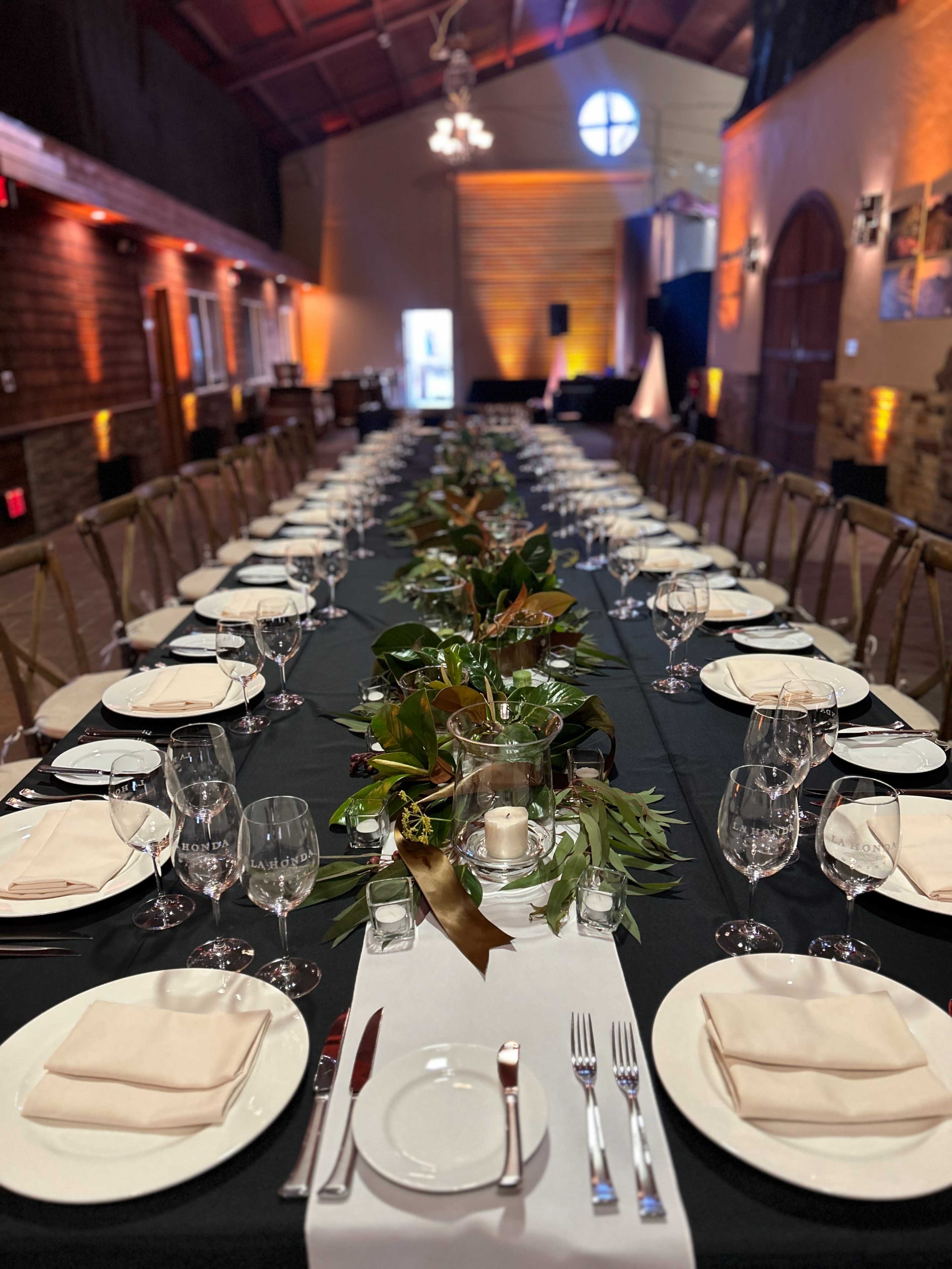A long, elegantly set dining table is arranged with white plates, glassware, and cloth napkins, featuring a central greenery centerpiece in a spacious venue.