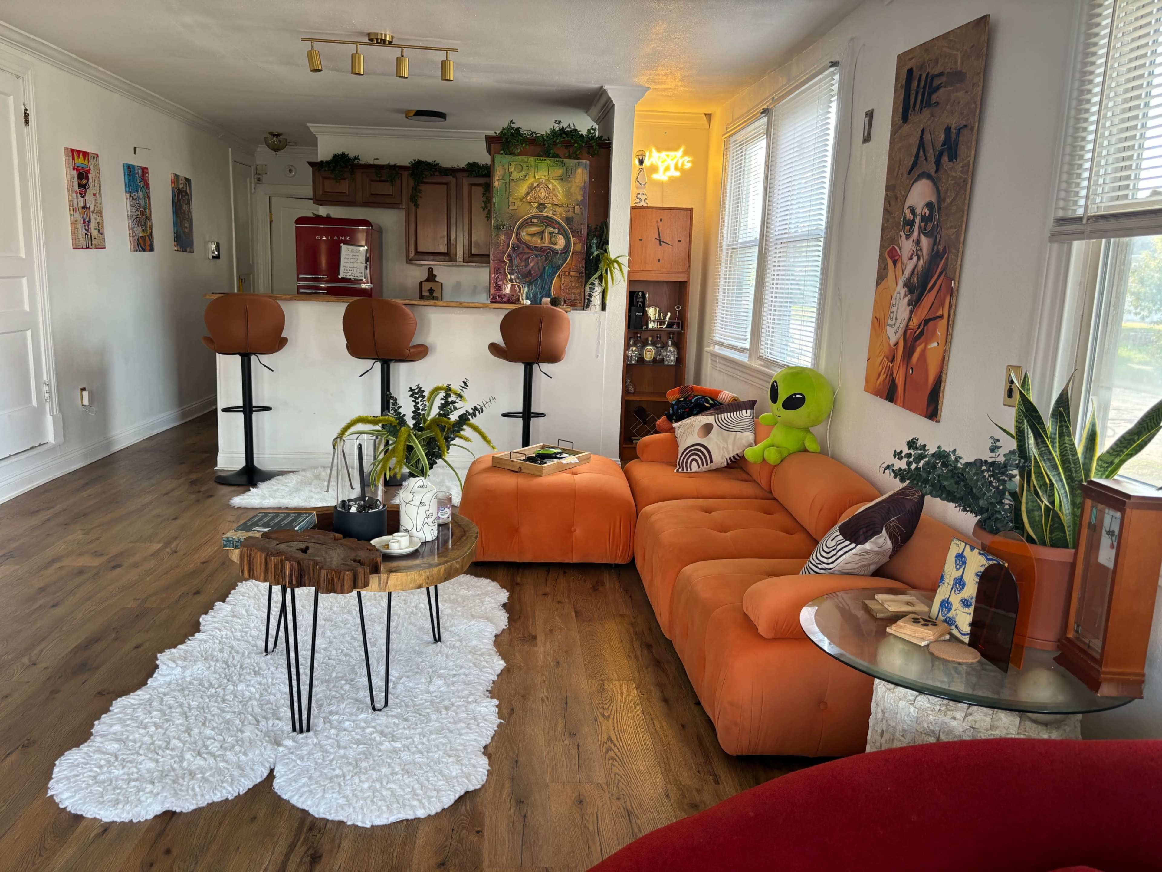 The living room features a vibrant orange sectional sofa, a round wooden coffee table on a white rug, and a bar area with three brown stools.