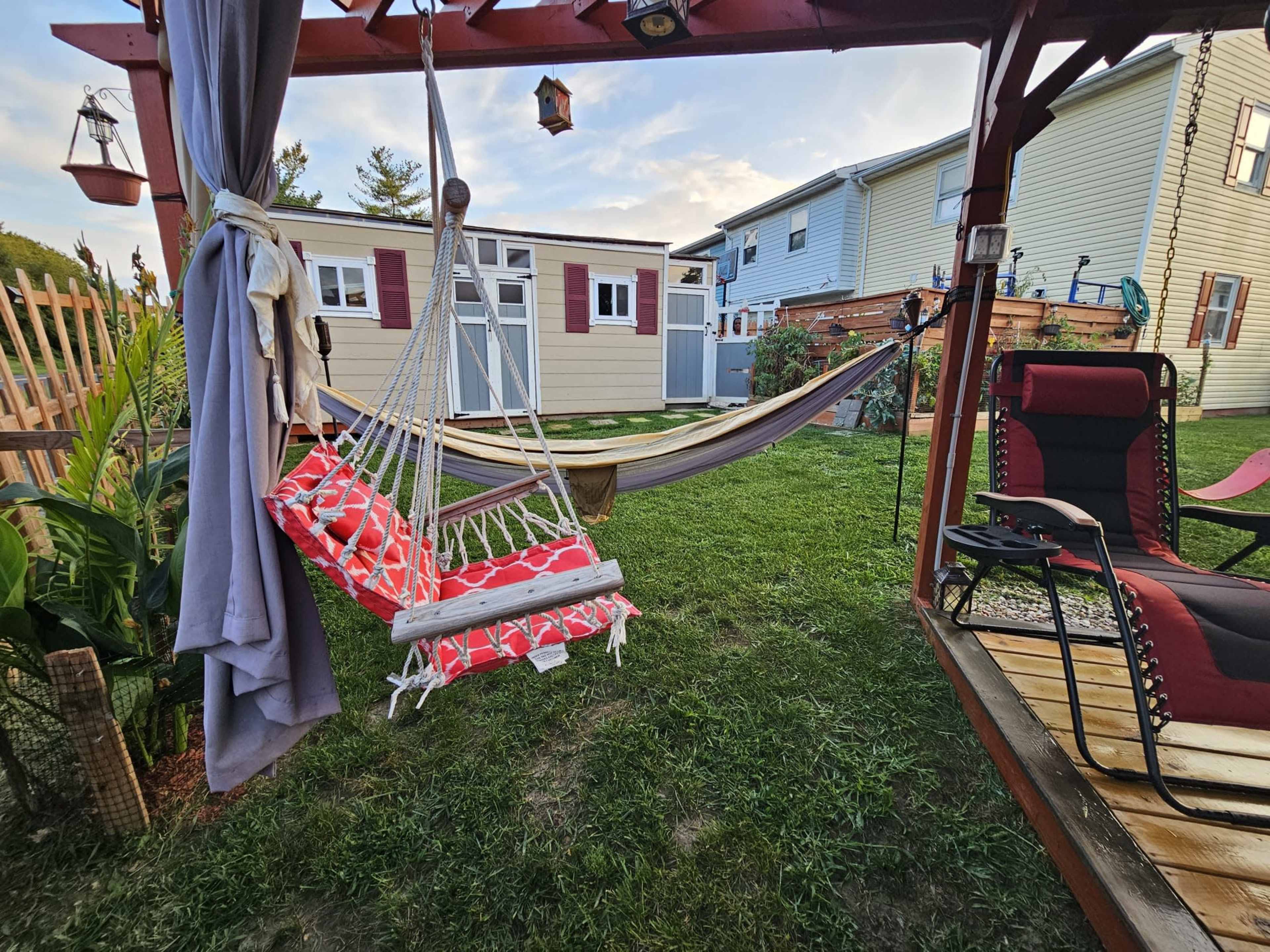 A backyard scene features a red and white hammock and a hanging chair under a wooden pergola, with a small shed and another hammock visible in the background.