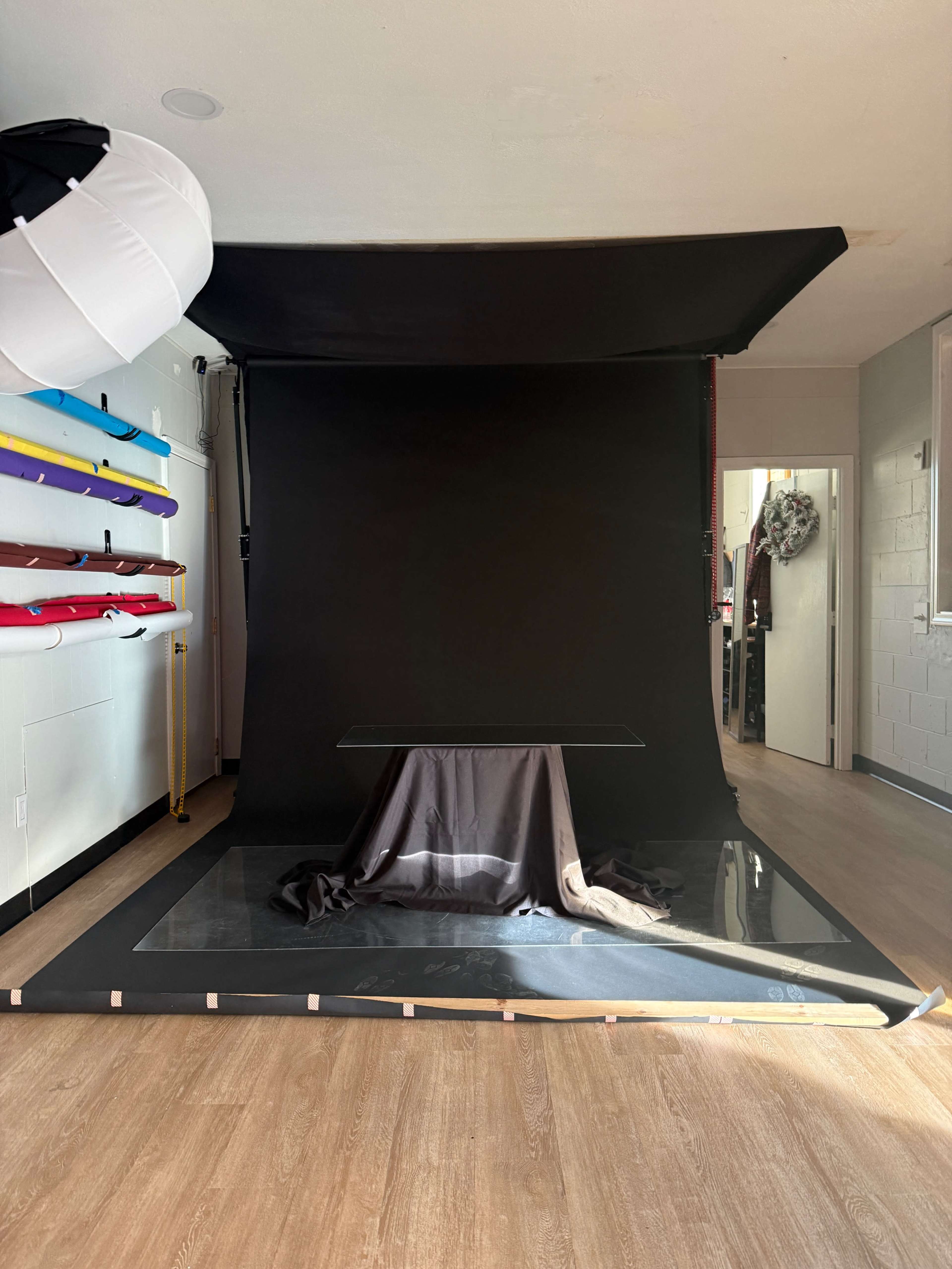 The image shows a photography studio featuring a black backdrop, a glass table with a draped cloth, and colorful equipment mounted on the wall.