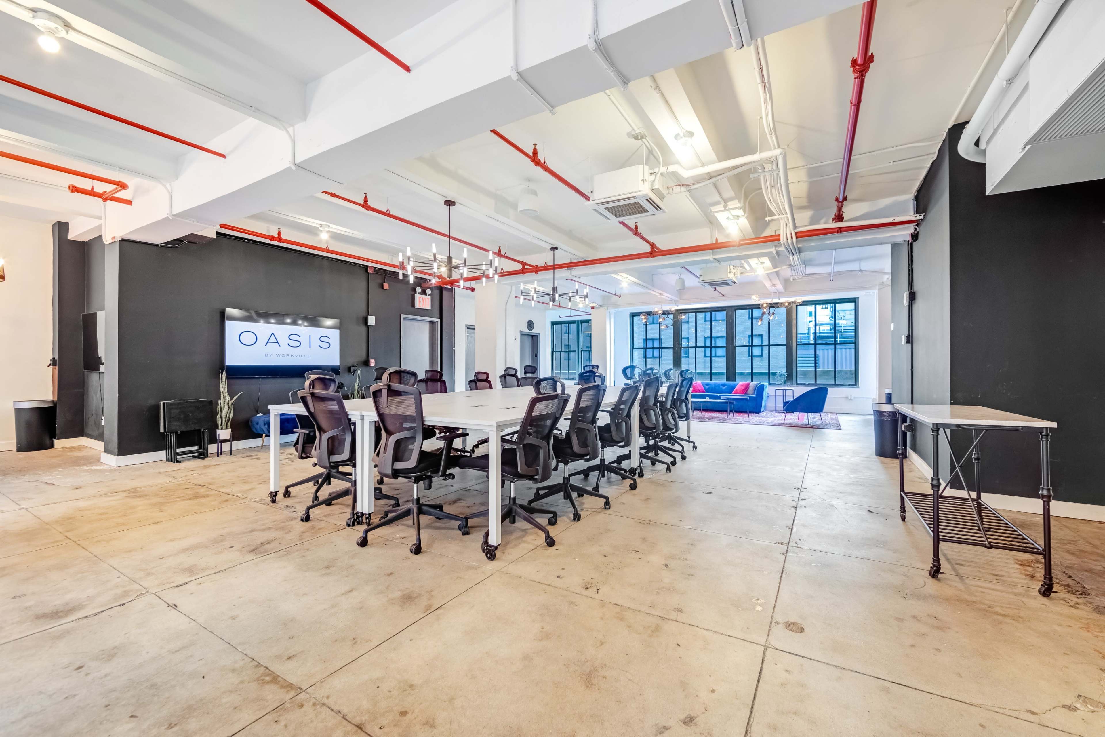 A modern conference room features a long table surrounded by ergonomic chairs, with a large screen displaying "OASIS" on one wall and a lounge area visible through the windows.