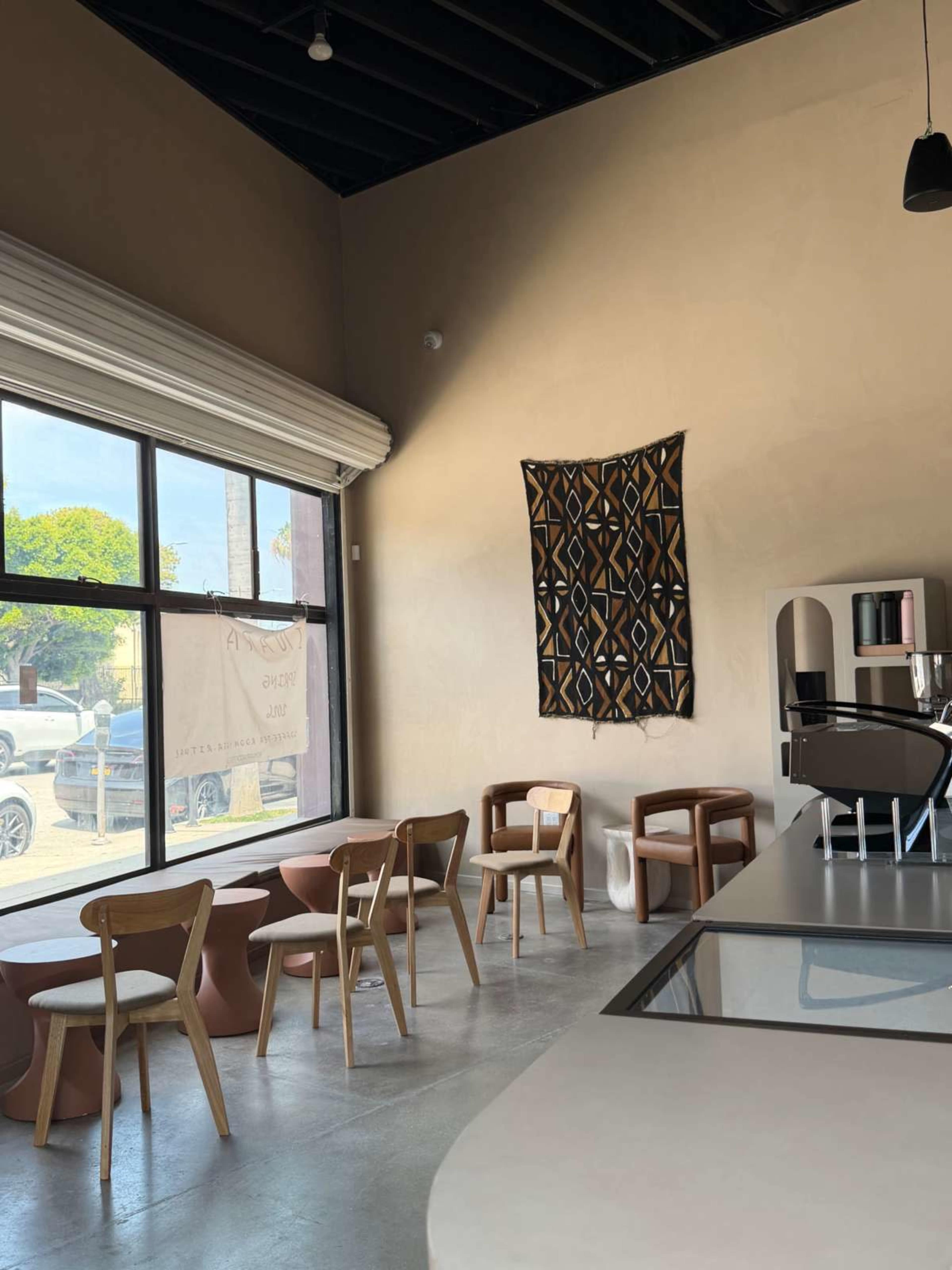 The interior of a café features wooden tables and chairs arranged around a minimalist space with a large window showing an outdoor view, and a patterned wall hanging.