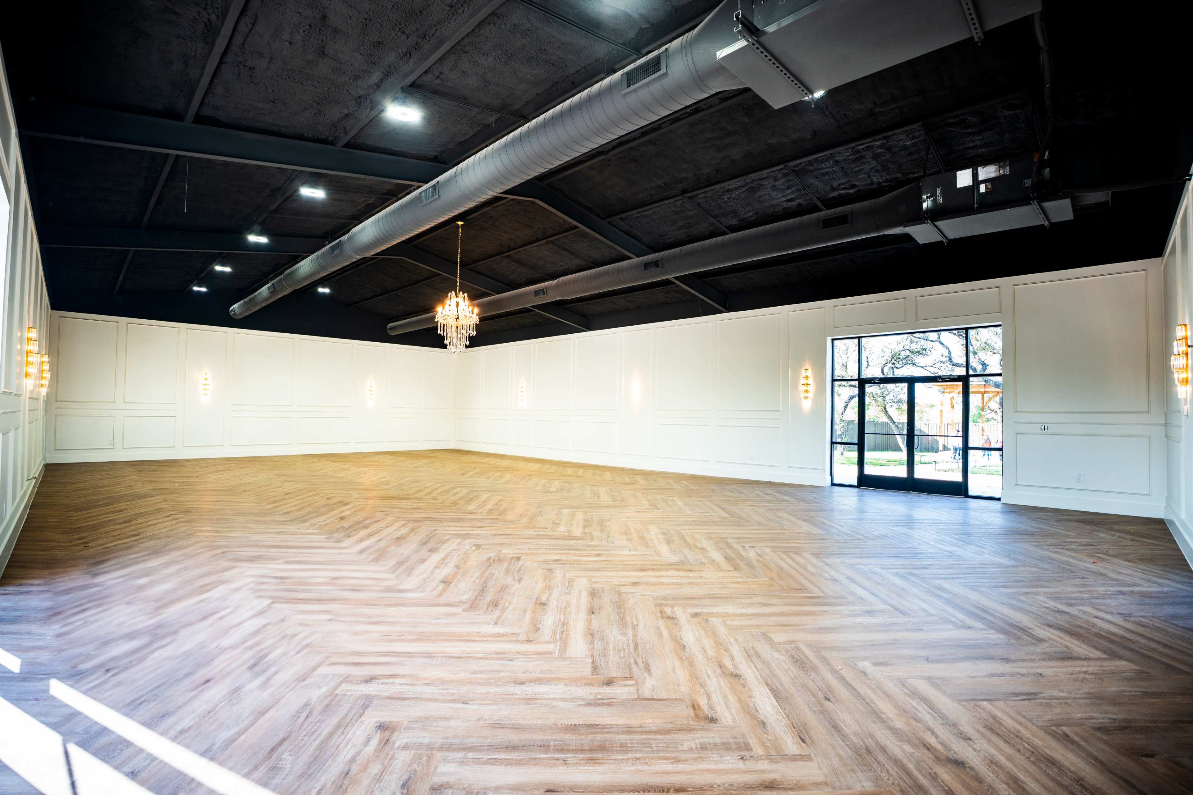 The image shows an empty hall with a wooden herringbone floor, white walls, and large windows allowing natural light to enter.