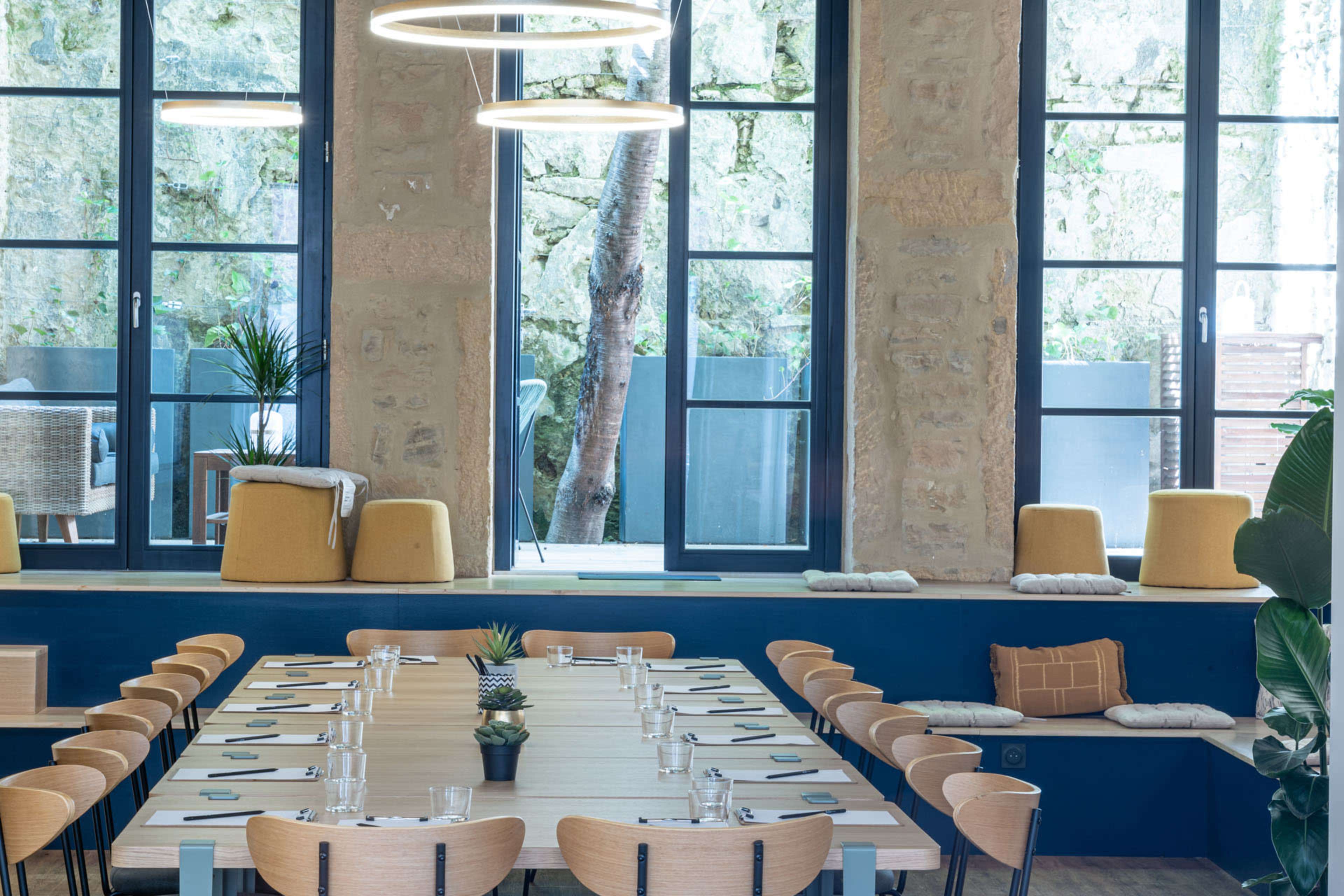 The image shows a modern dining space with a large wooden table set for a meal, surrounded by wooden chairs, and large windows letting in natural light.