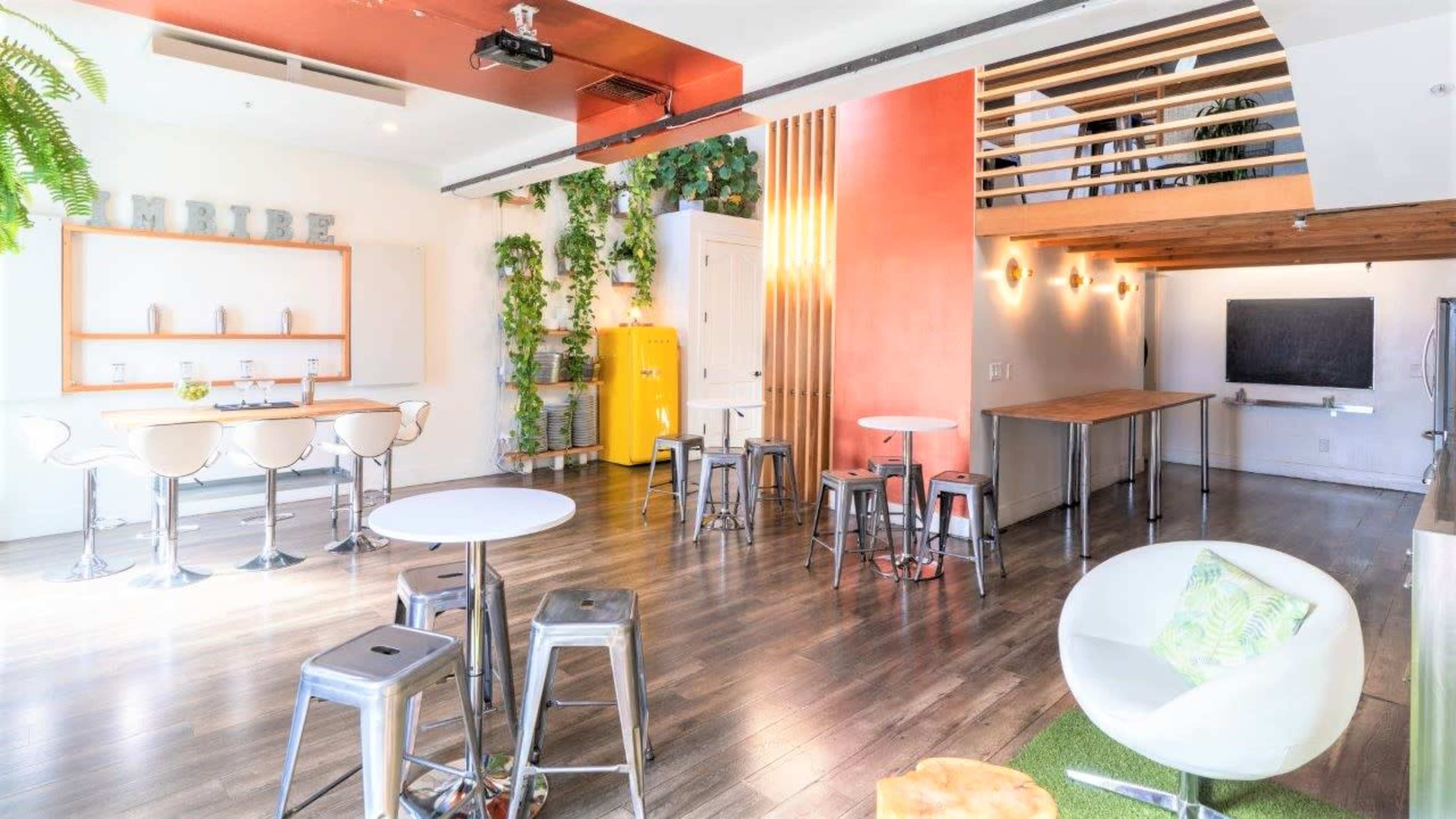 The image shows a modern, open interior space with high ceilings, featuring wooden floors, white walls, and a mix of seating options, including bar stools and tables.