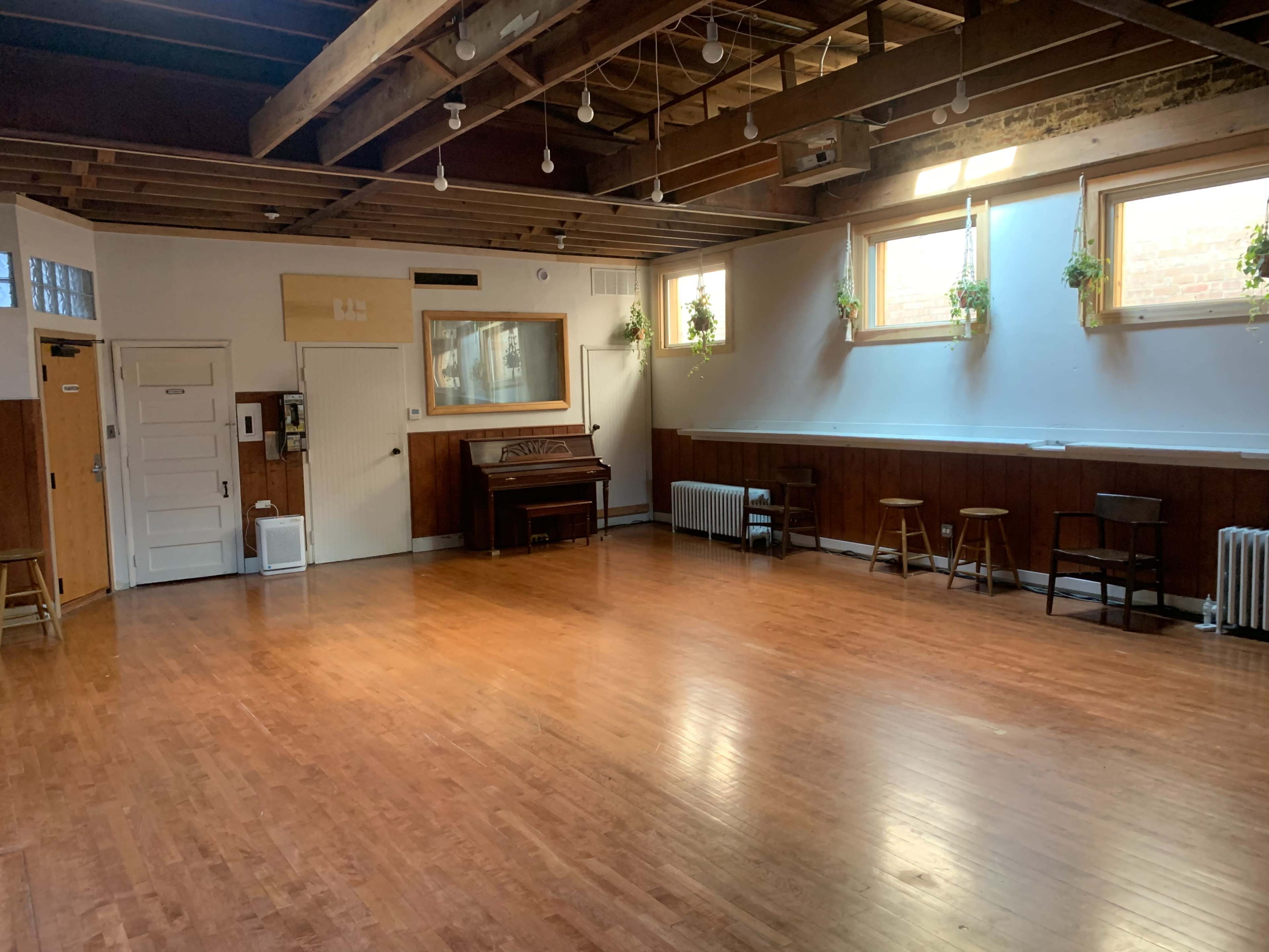 The image shows a spacious, empty wooden-floored room with tall windows, a piano against one wall, and a few chairs arranged along another wall.