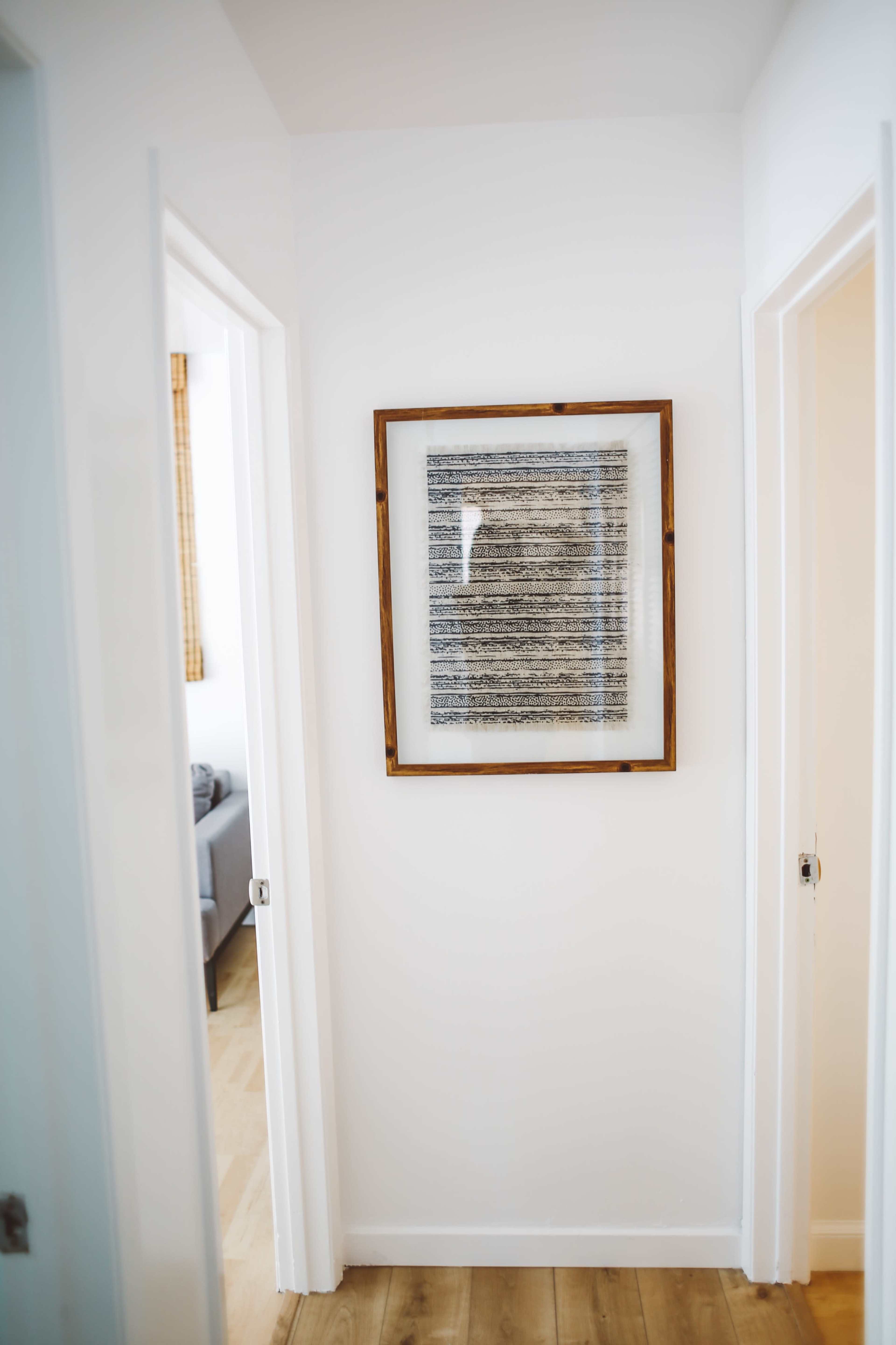 A framed abstract artwork hangs on a white wall in a narrow hallway with two open doorways on either side.