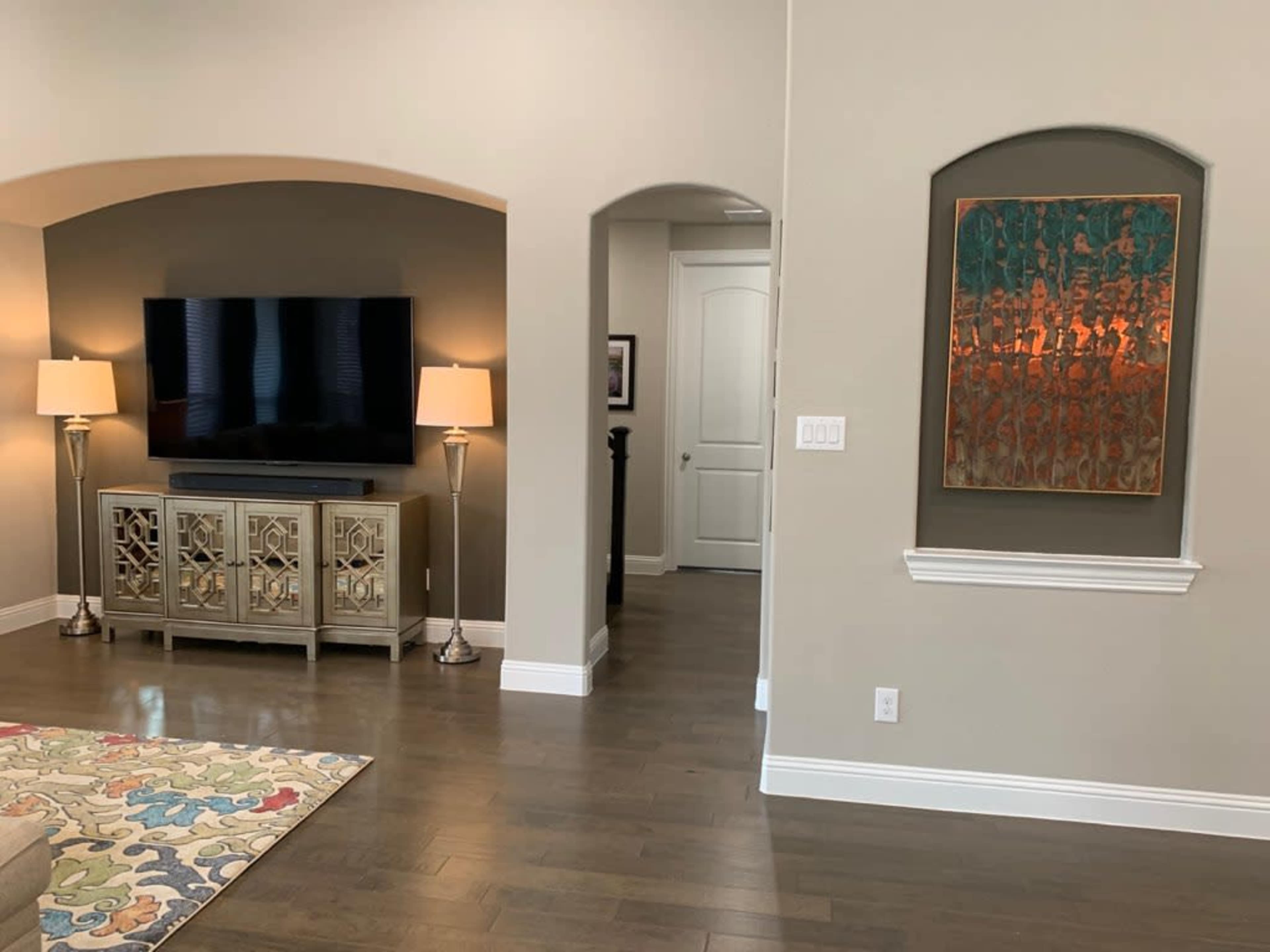 A living room with a large wall-mounted TV, two floor lamps, a decorative console table, and an abstract artwork on the wall, leading to a hallway with a closed door.
