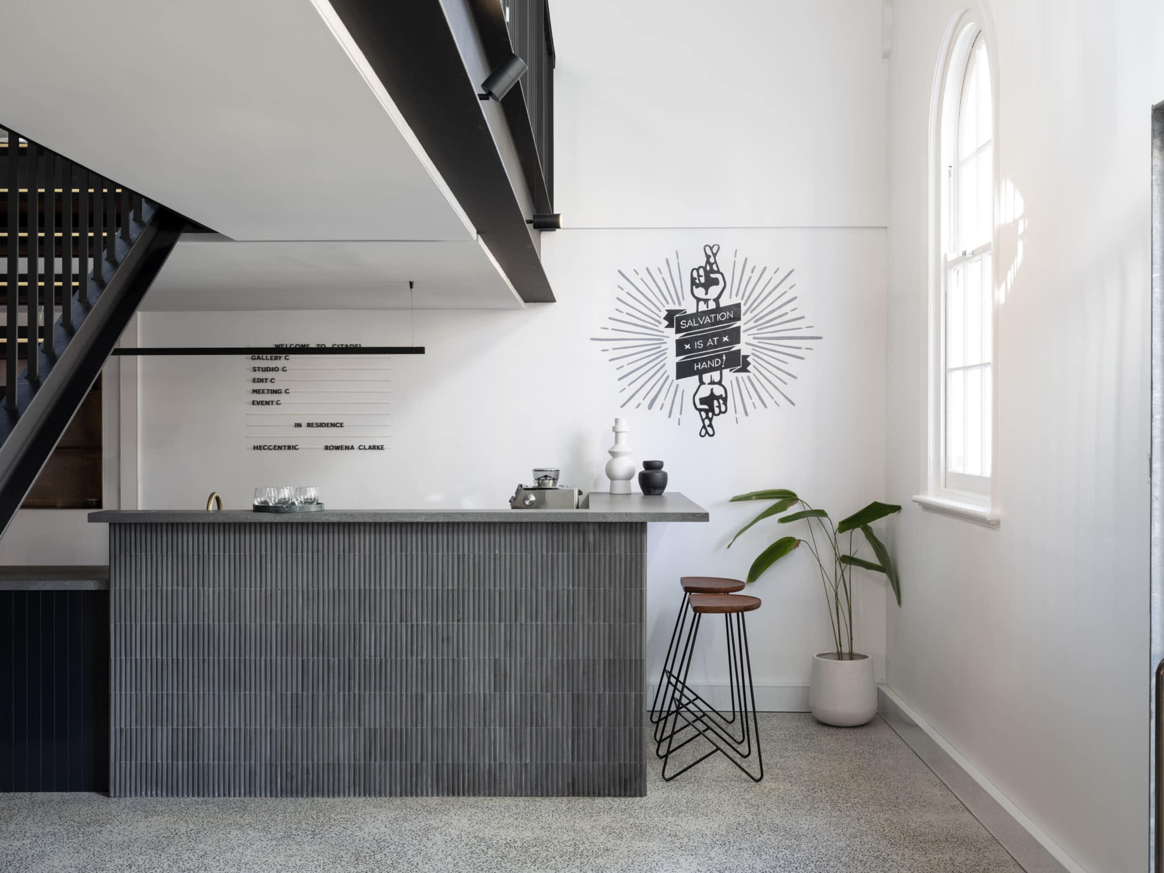 A modern reception area features a sleek gray counter with vertical stripes, a minimalist decoration on the wall, and a potted plant beside a bar stool.