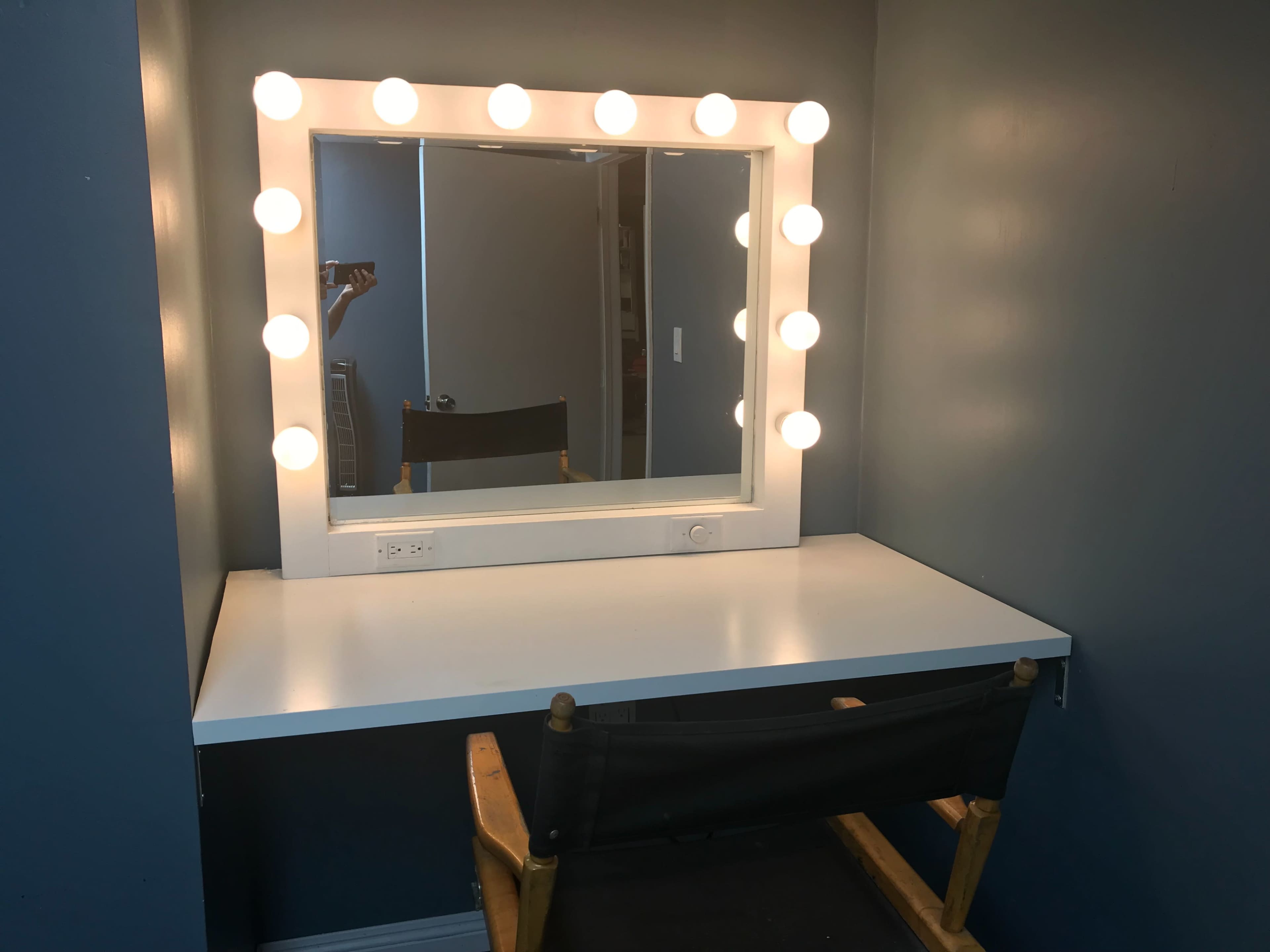 A makeup vanity with a large lighted mirror, a white countertop, and a black director's chair in front of it against a gray wall.
