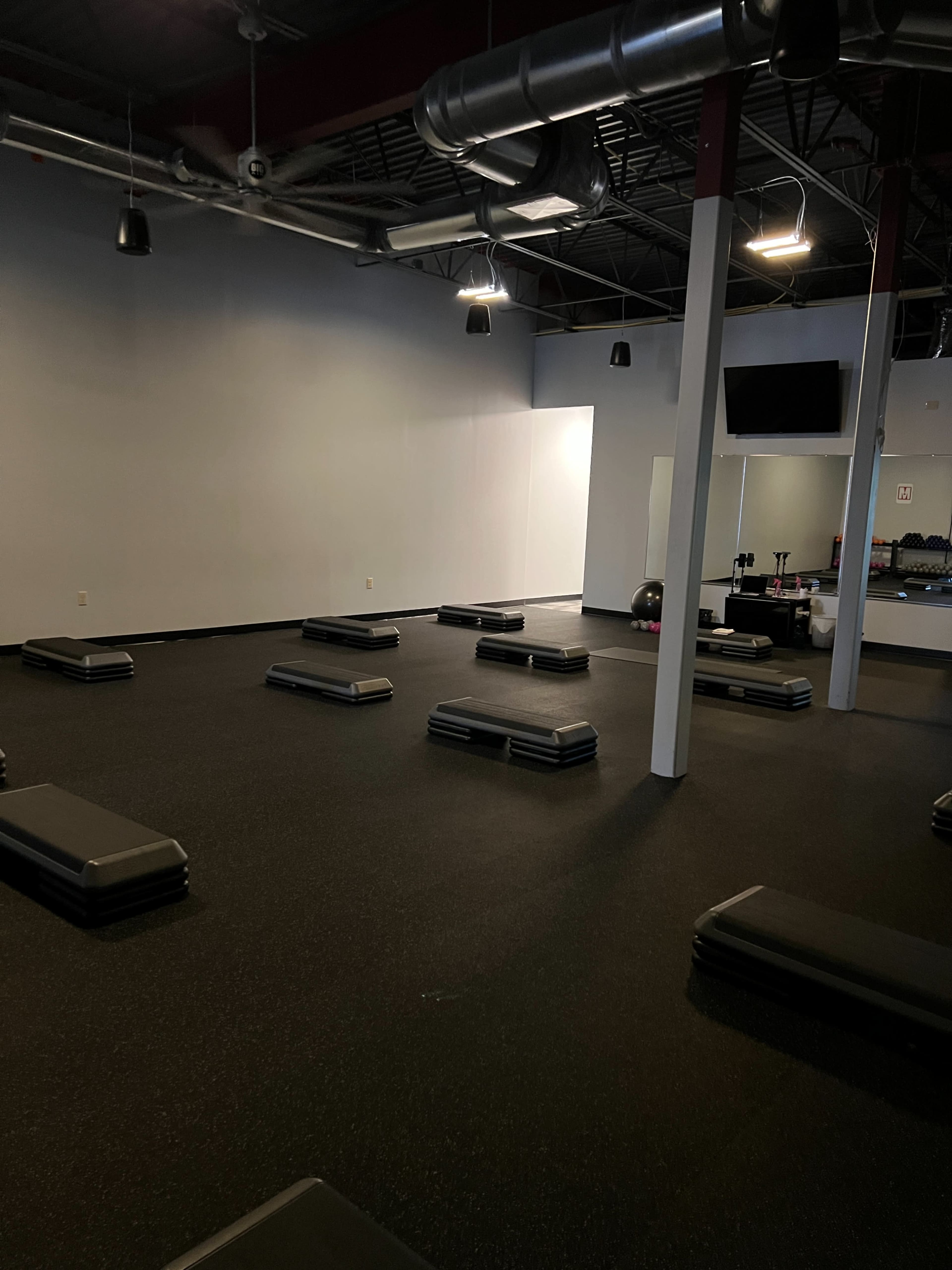 A spacious gym studio with several step platforms arranged on a rubber floor, under exposed ductwork and overhead lighting.