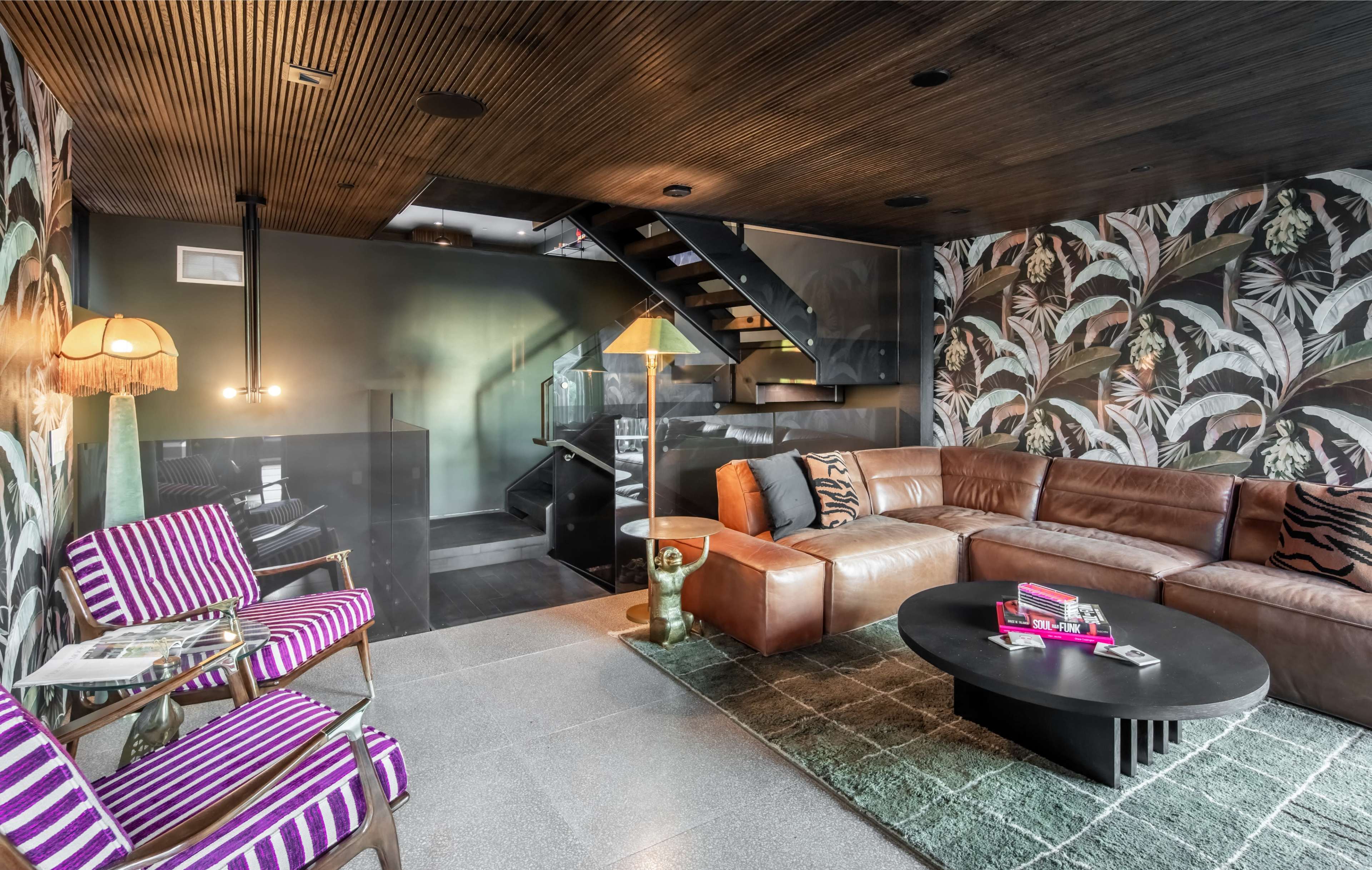 A modern living room featuring a sleek leather sectional sofa, a round coffee table, purple-striped chairs, and a staircase in the background, all framed by vibrant wallpaper and a mix of decorative lighting.