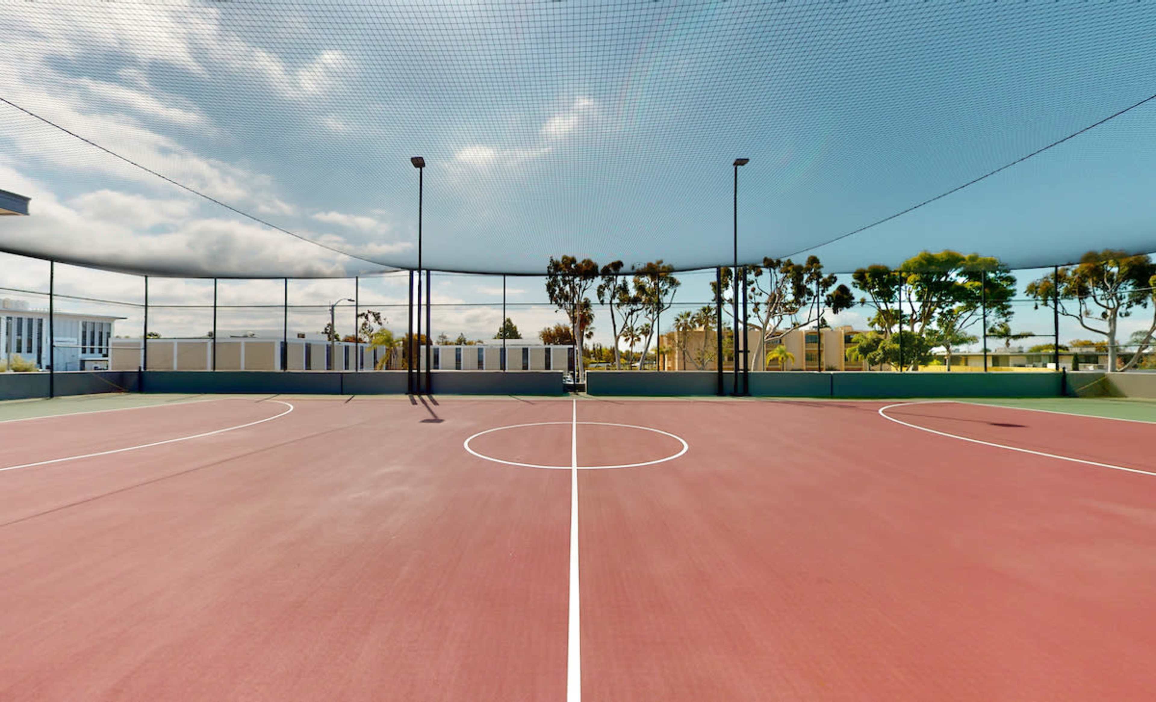 An empty outdoor basketball court surrounded by a net and trees under a cloudy sky.