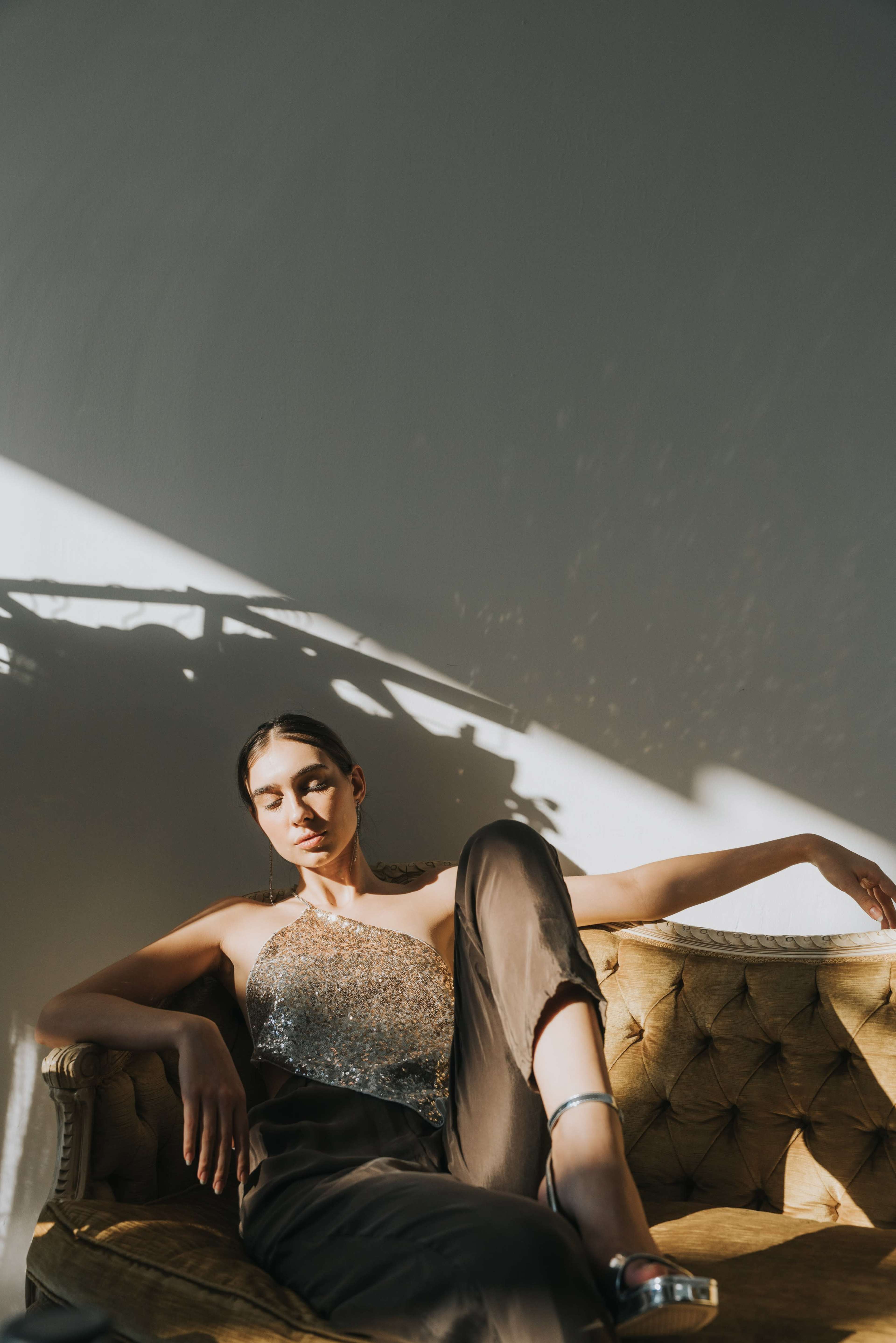 A woman in a sparkling top sits on a vintage couch, casting shadows against a wall as sunlight filters through.